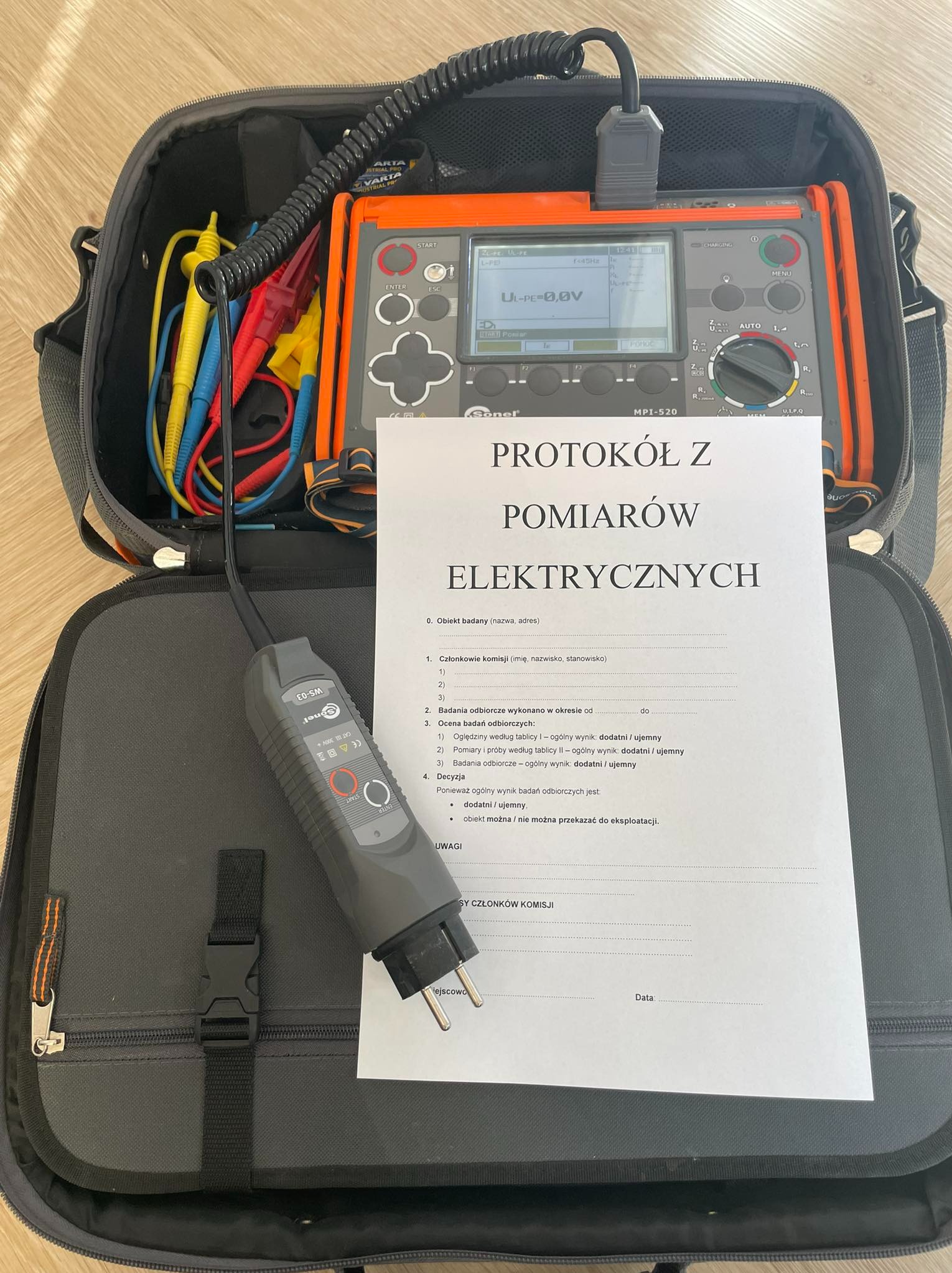 Protokół z pomiarów elektrycznych leżący obok miernika Sonel MPI-520 w torbie. Widoczne przewody pomiarowe i wtyczka. Dokument częściowo zasłania sprzęt.