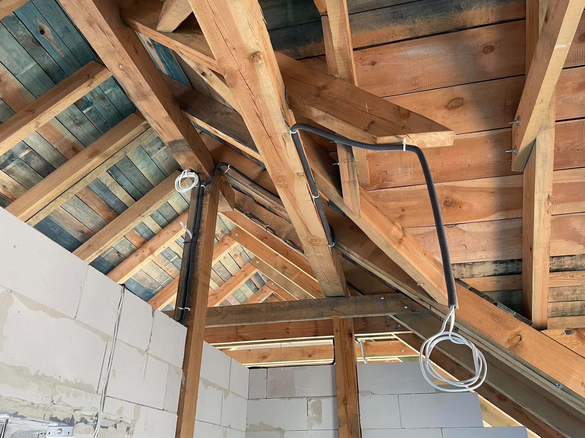 Instalacja elektryczna w budynku w trakcie budowy. Widoczne kable prowadzone po drewnianej konstrukcji dachu i ścianach z bloczków. Kable zabezpieczone peszlami.