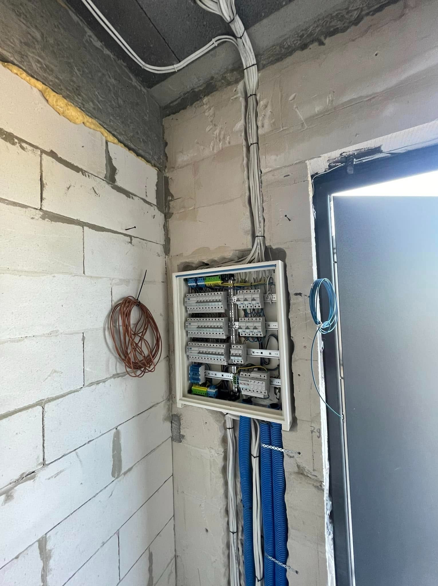 Rozdzielnia elektryczna w surowym wnętrzu, widoczne kable w peszlach i przewody na ścianie z bloczków. Instalacja w trakcie realizacji.