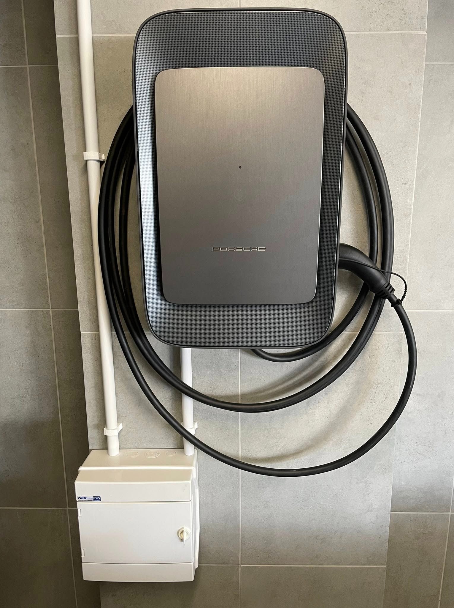 Zamontowana na ścianie ładowarka do samochodu elektrycznego Porsche, z kablem i skrzynką elektryczną. Minimalistyczny design, szare płytki w tle.