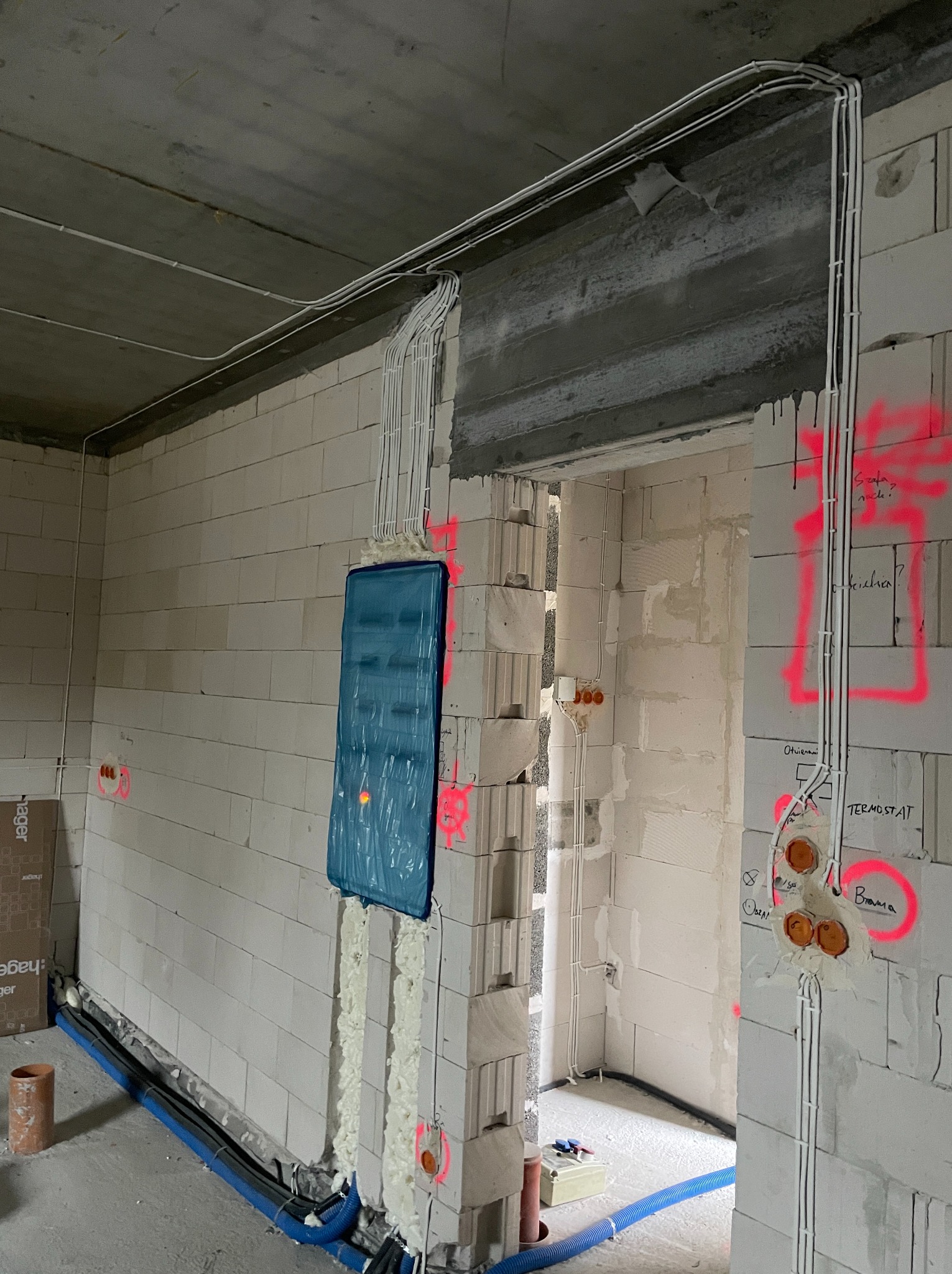 Białe przewody elektryczne prowadzone po ścianach z betonu komórkowego w surowym wnętrzu. Otwory na gniazdka i włączniki zaznaczone różowym sprayem.