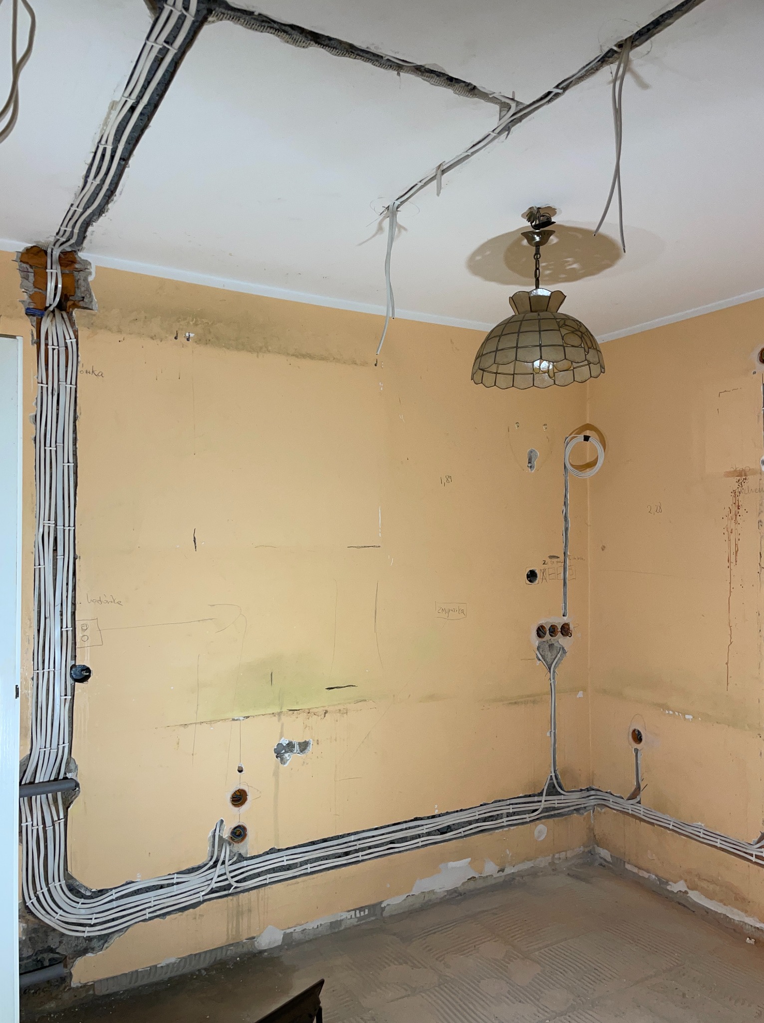 Wnętrze w trakcie remontu z widocznymi przewodami elektrycznymi ułożonymi na ścianach i suficie. Widoczne puszki i punkty podłączeń. Stara lampa sufitowa.