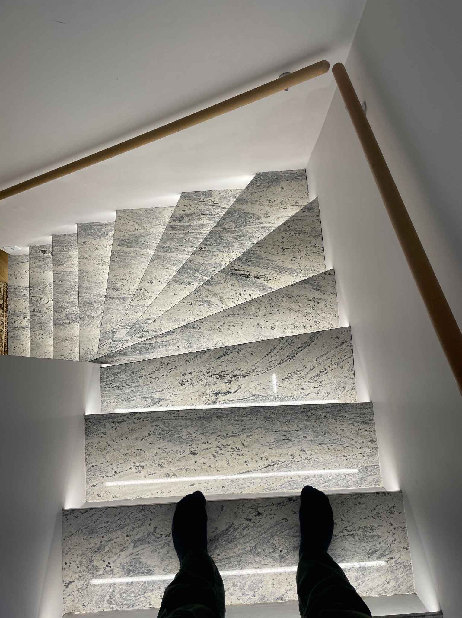 Nowoczesne schody z podświetleniem LED, widok z góry na marmurowe stopnie i drewnianą poręcz. U dołu kadru widoczne stopy osoby stojącej na schodach.
