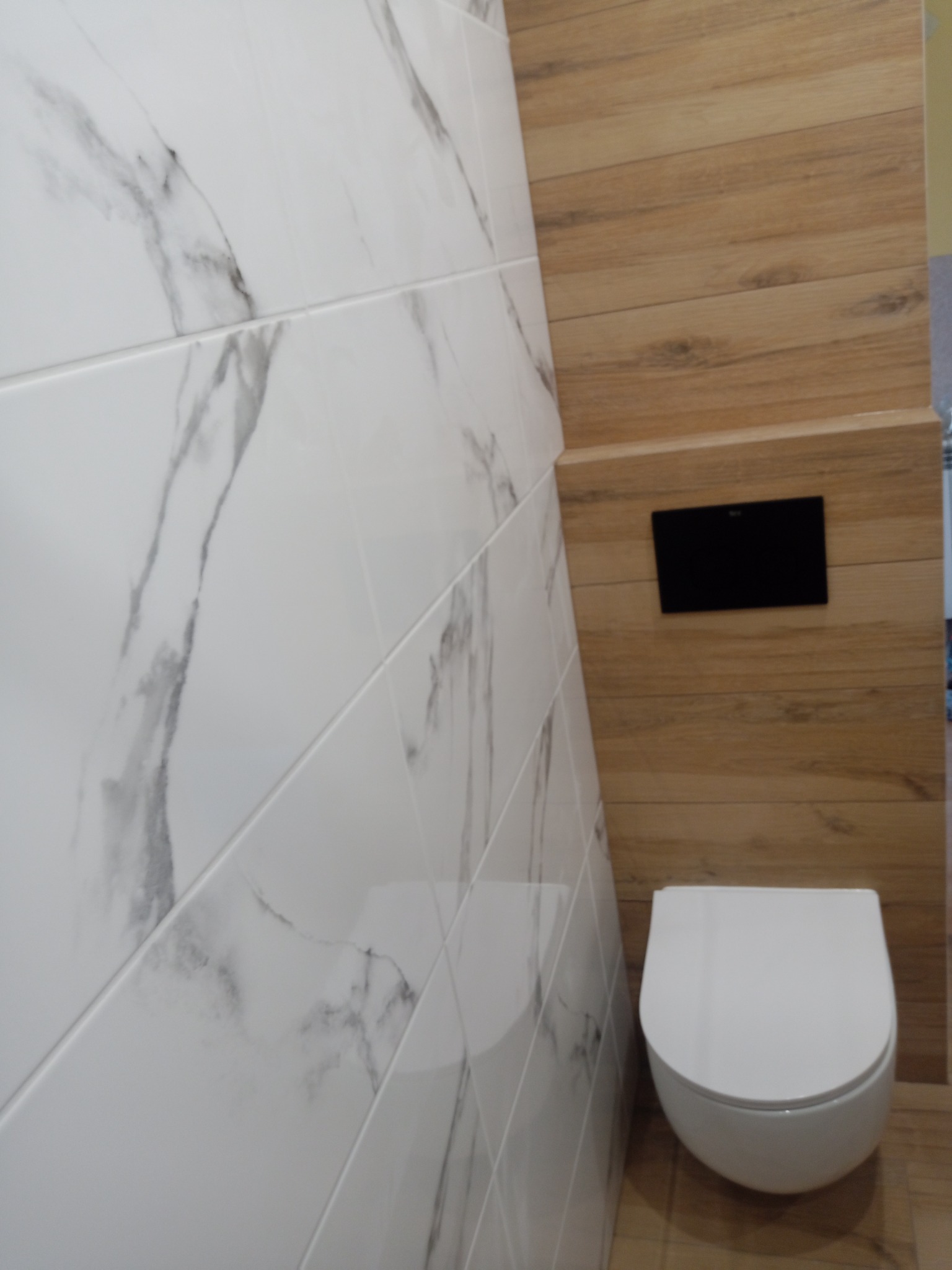 Nowoczesna toaleta podwieszana z białym sedesem i czarnym przyciskiem spłukującym, na tle ściany wyłożonej płytkami imitującymi drewno i marmur.