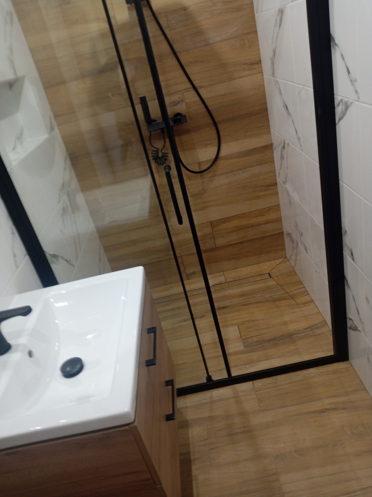 Nowoczesna łazienka z kabiną prysznicową typu walk-in, drewnopodobnymi płytkami na podłodze i ścianie, oraz białą umywalką z szafką w kolorze drewna.