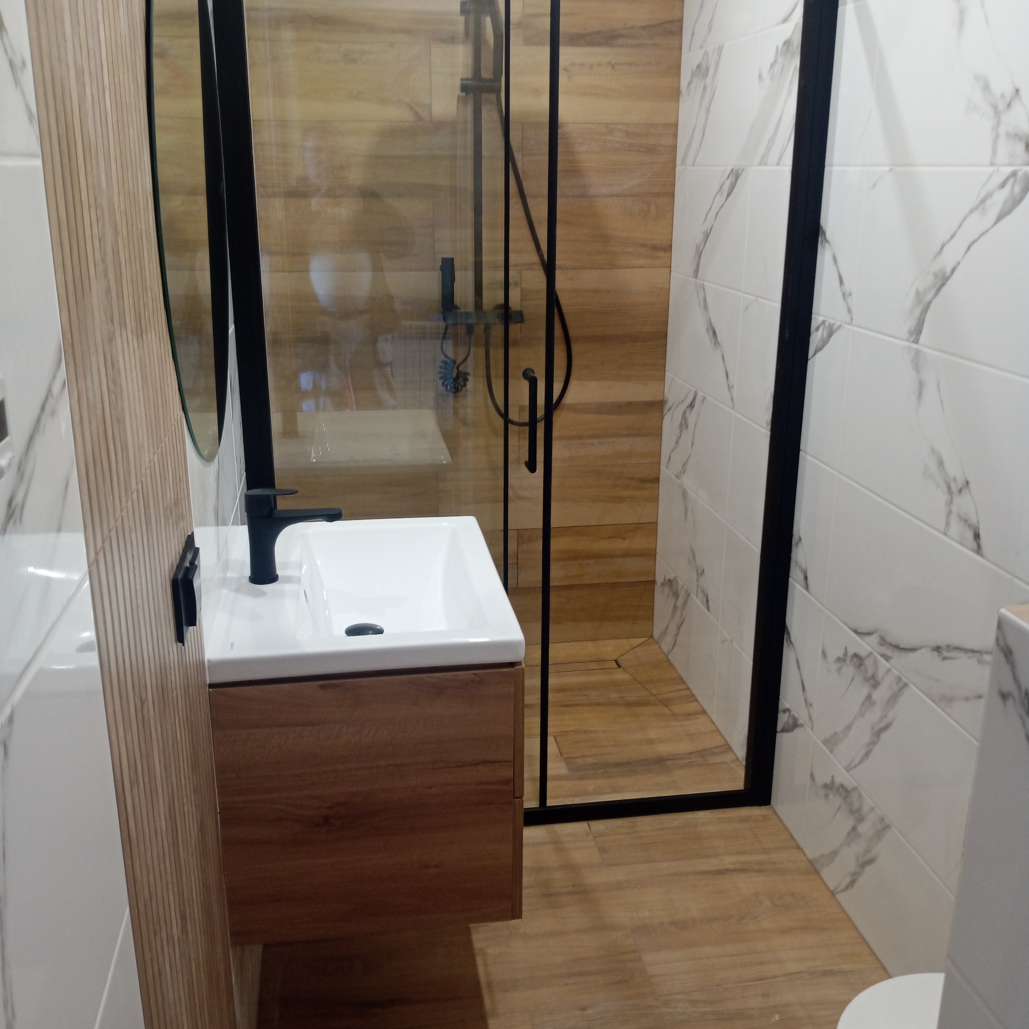 Nowoczesna łazienka z kabiną prysznicową typu walk-in, umywalką nablatową na drewnianej szafce i białymi płytkami imitującymi marmur. Czarna armatura i rama kabiny.