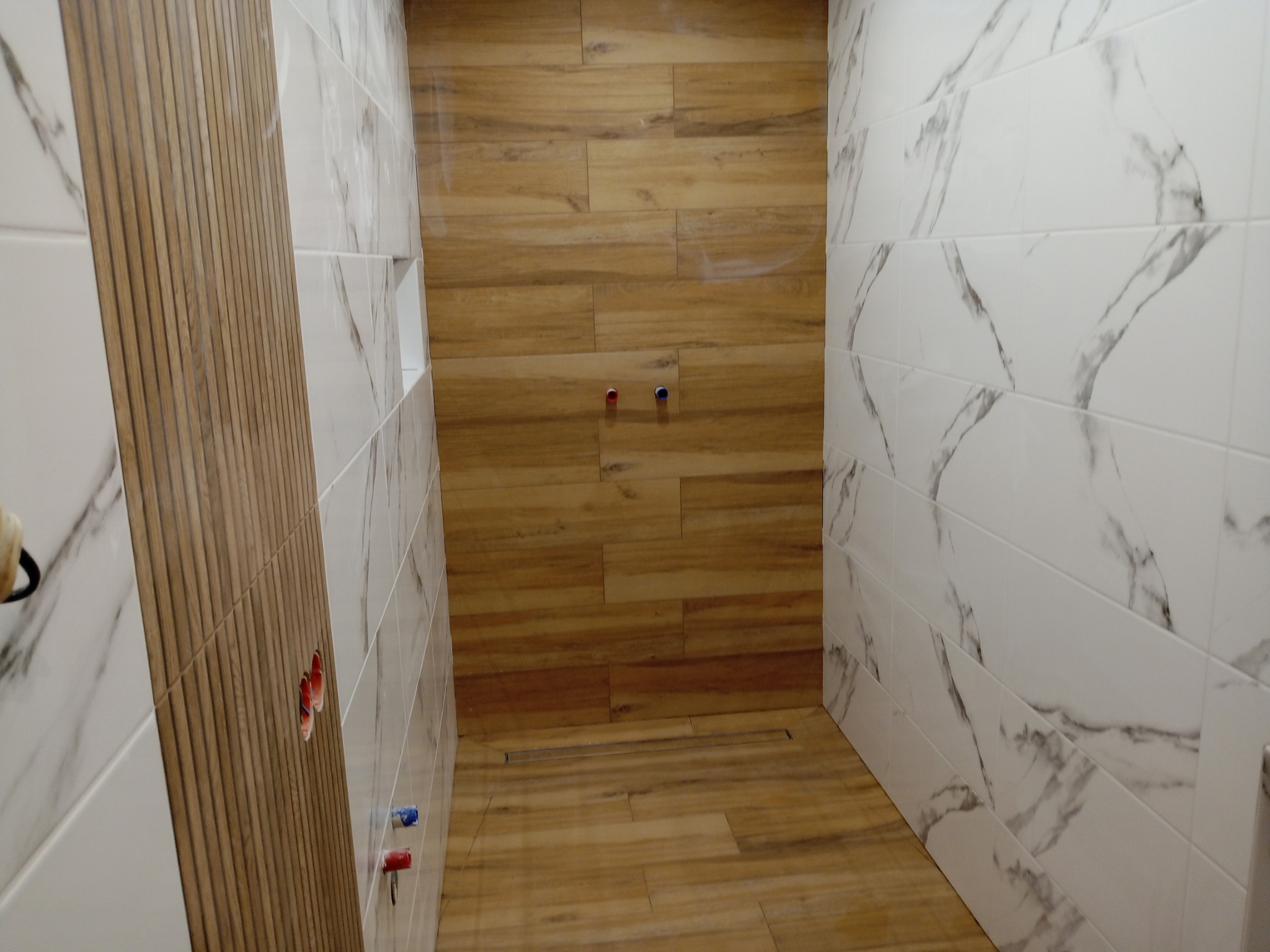 Wykończona kabina prysznicowa z drewnopodobnymi i marmurowymi płytkami. Widoczne odpływ liniowy i wystające zawory. Ściana z dekoracyjnymi panelami.