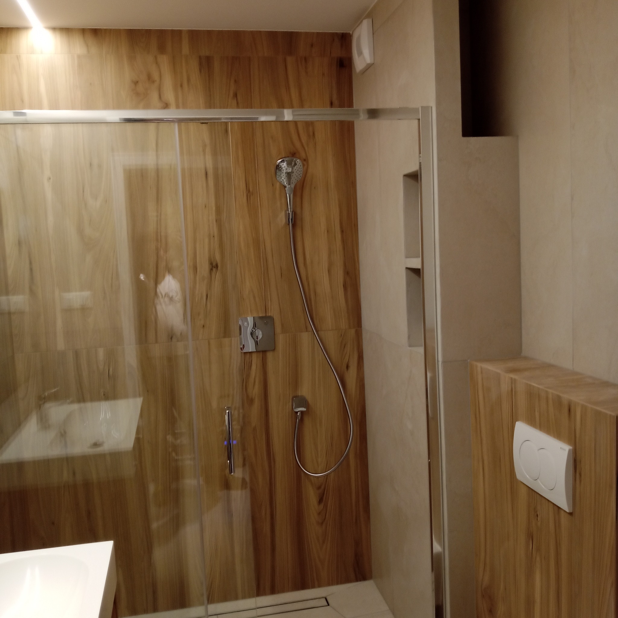 Nowoczesna kabina prysznicowa z drewnopodobnymi płytkami, szklane drzwi przesuwne, minimalistyczna armatura i ukryta spłuczka. Jasne, ciepłe oświetlenie.