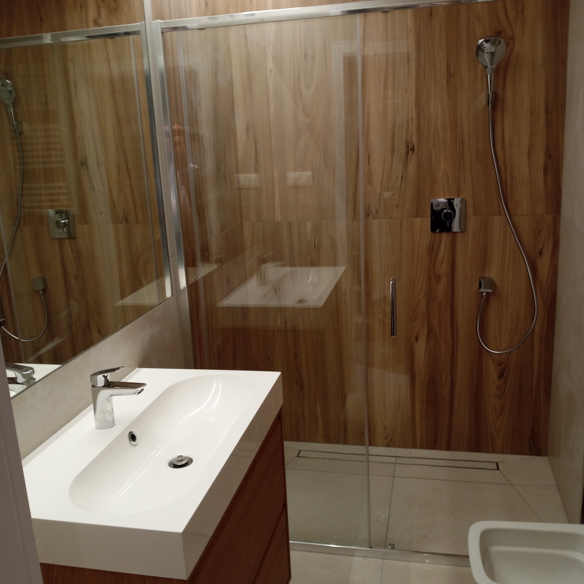 Nowoczesna łazienka z kabiną prysznicową typu walk-in, drewnopodobnymi płytkami na ścianach i białą umywalką nablatową z szafką w kolorze drewna.