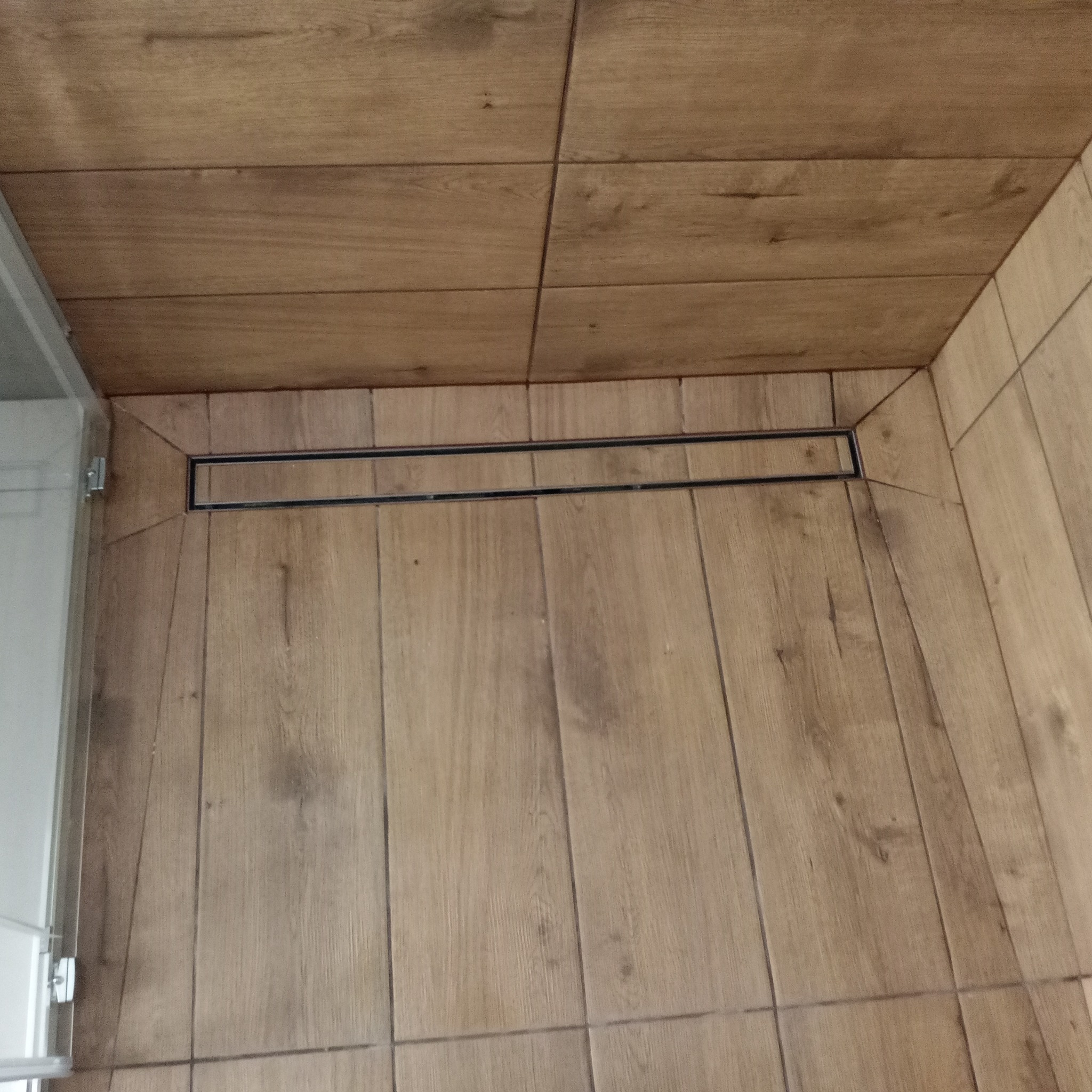 Ukończona kabina prysznicowa z odpływem liniowym i płytkami imitującymi drewno na ścianach i podłodze. Widoczny fragment szklanej ściany kabiny.