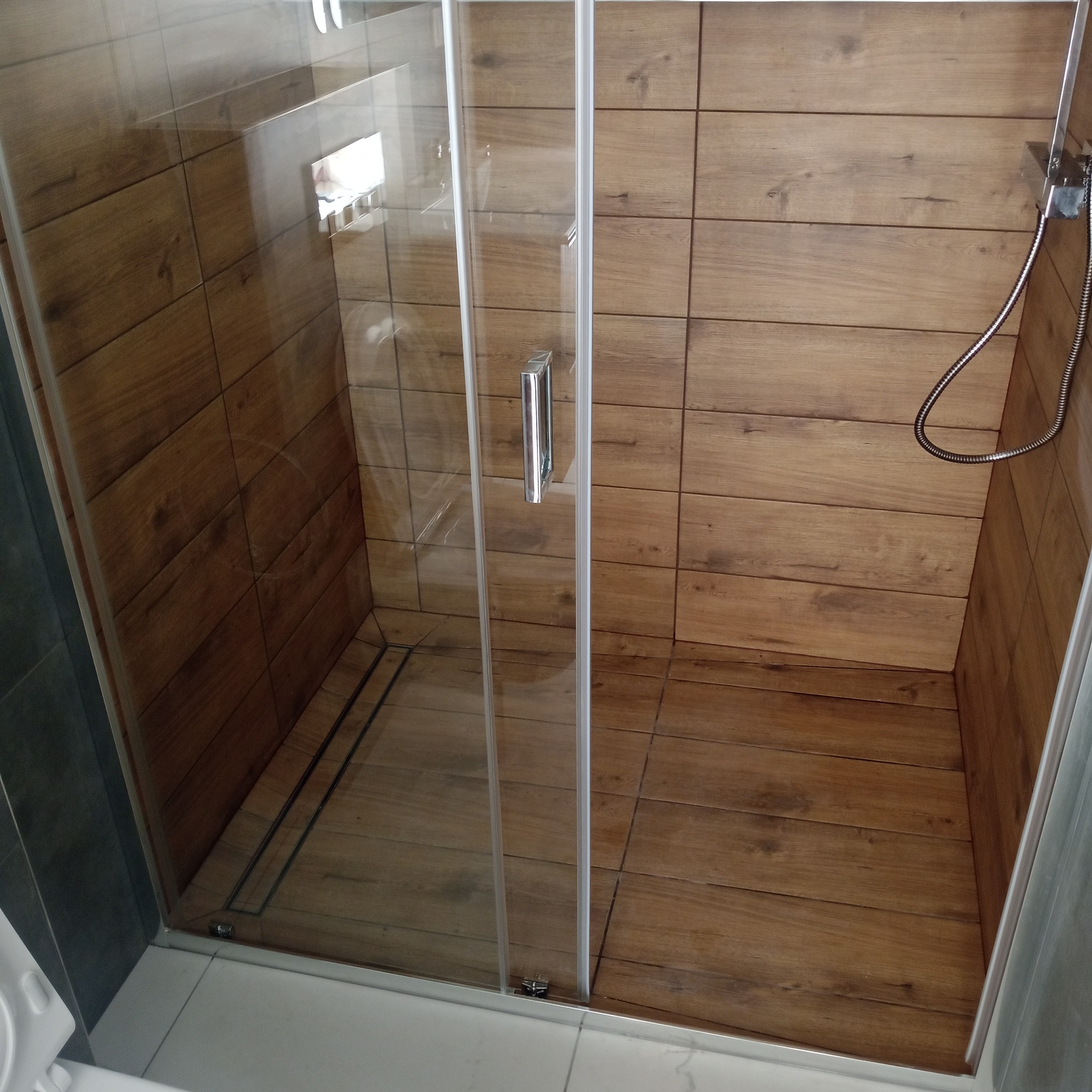Kabina prysznicowa z drewnopodobnymi płytkami, chromowana armatura i odpływ liniowy. Nowoczesny design, jasne wnętrze.