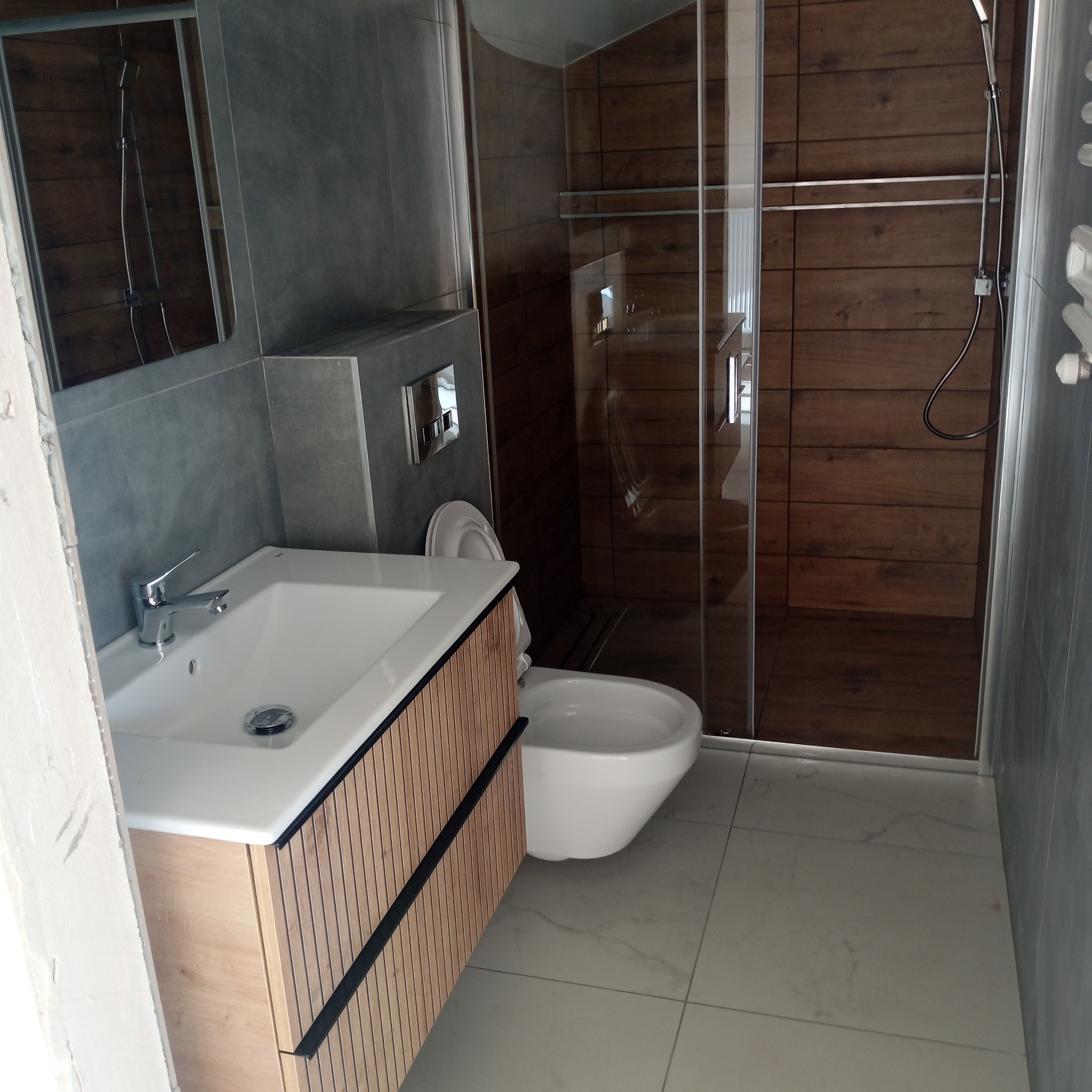 Nowoczesna łazienka po remoncie: szare płytki, drewnopodobna kabina prysznicowa, biała umywalka z szafką z lamelami i toaleta. Marmurowe płytki podłogowe.