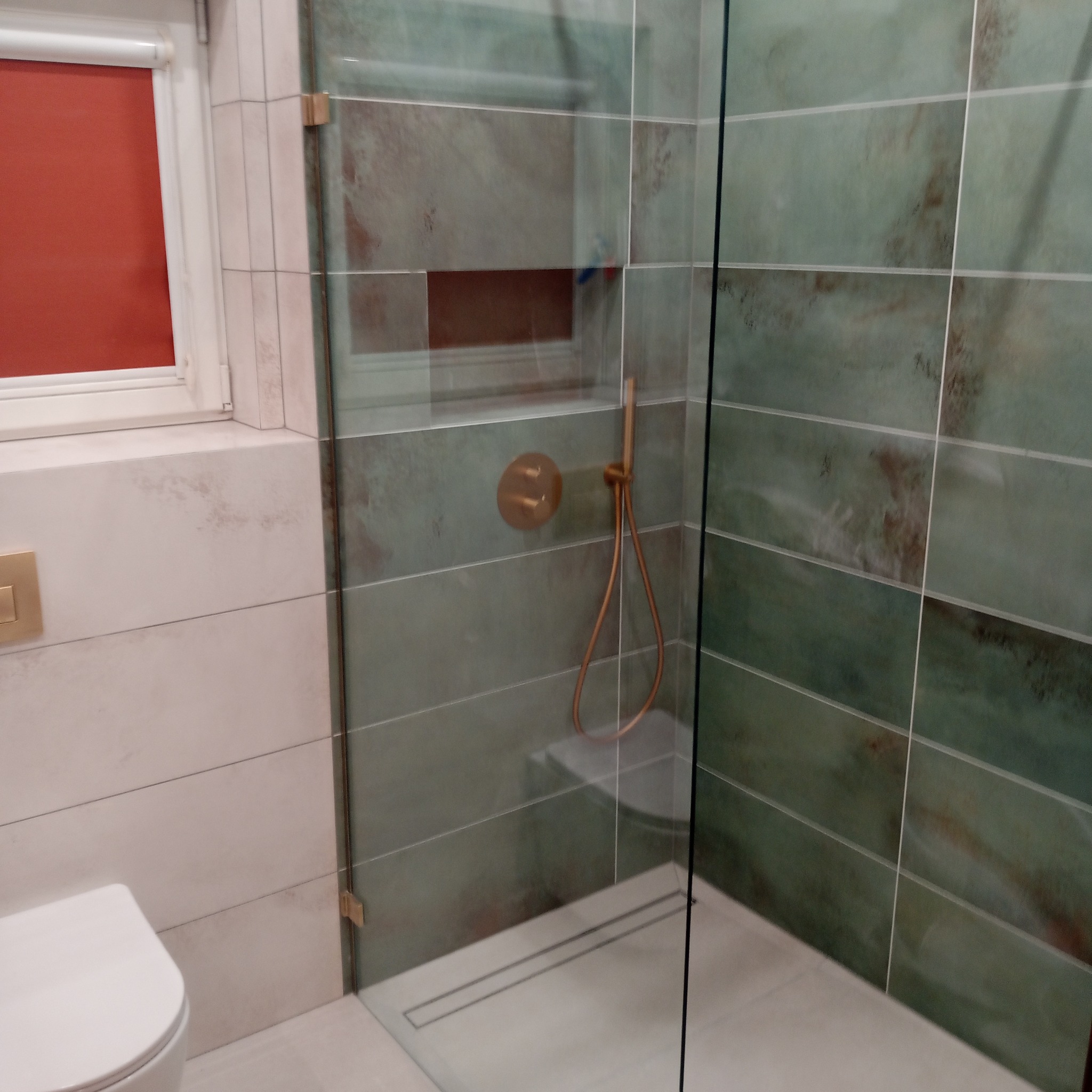 Nowoczesna kabina prysznicowa z zielonymi płytkami imitującymi kamień, złotą armaturą i odpływem liniowym. Widoczny fragment toalety i okno z czerwoną roletą.