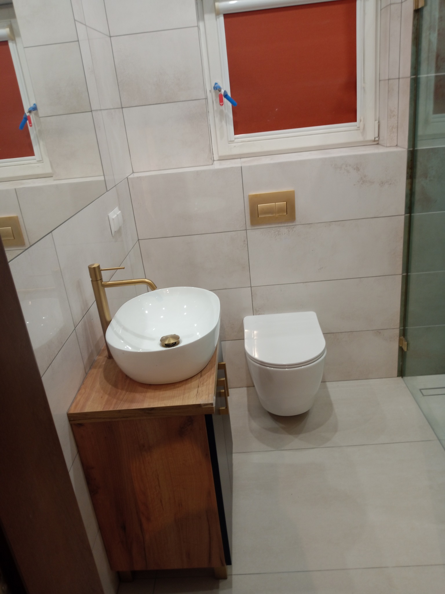 Nowoczesna łazienka z umywalką nablatową na drewnianej szafce, wiszącą toaletą i beżowymi płytkami. Złota armatura i detale dodają elegancji.