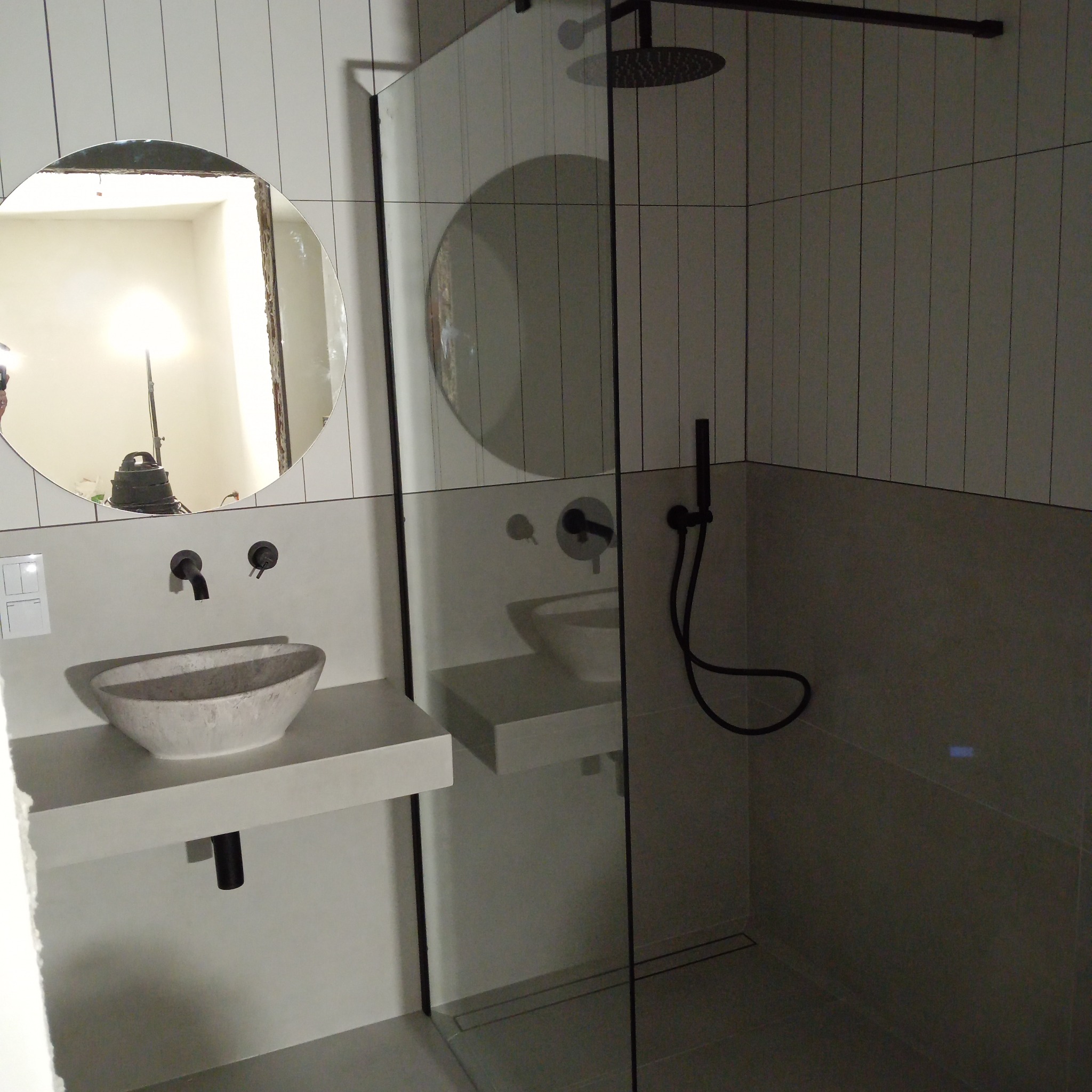 Nowoczesna łazienka z owalnym lustrem, kamienną umywalką i kabiną prysznicową typu walk-in z czarnymi detalami. Minimalistyczny design, jasne płytki.