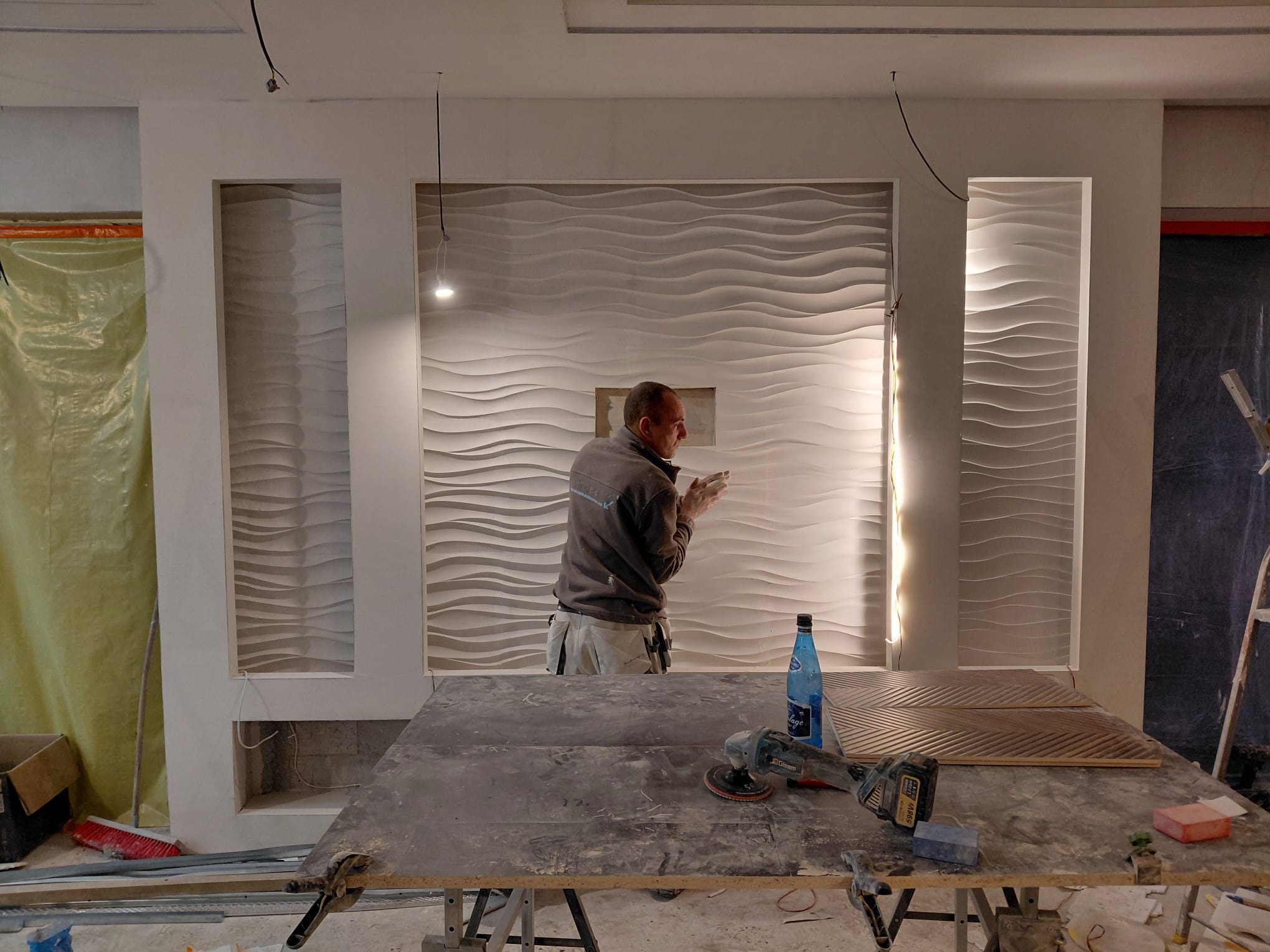 Montaż dekoracyjnych paneli ściennych 3D w salonie. Widoczny pracownik podczas pracy, narzędzia na stole i fragmenty paneli. Oświetlenie punktowe podkreśla fakturę.