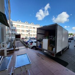 Pakuś Przeprowadzki - Otwarty furgon załadowany meblami i kartonami, zaparkowany przed budynkiem. Widoczne akcesoria do pakowania i transportu. Błękitne niebo z chmurami w tle.