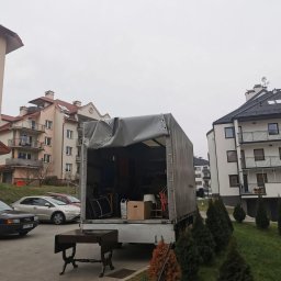 Pakuś Przeprowadzki - Otwarta paka samochodu dostawczego załadowana meblami i kartonami na tle budynków mieszkalnych. Na pierwszym planie stół z ciemnego drewna.