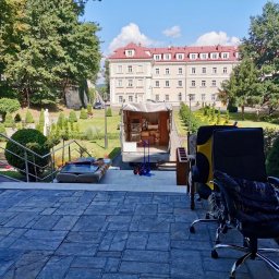 Pakuś Przeprowadzki - Przeprowadzka z eleganckiego budynku: meble biurowe i sofa na zewnątrz, w tle otwarty samochód dostawczy i budynek z czerwonym dachem w otoczeniu zieleni.
