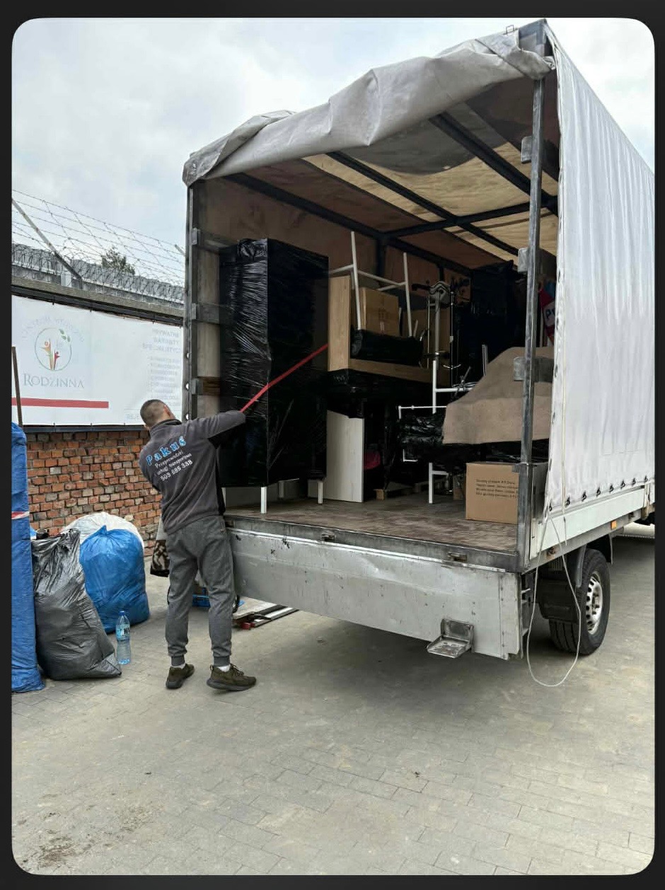 Pracownik firmy transportowej zabezpiecza meble w busie z otwartą plandeką. Widoczne zapakowane szafy, krzesła i inne przedmioty gotowe do przewozu.