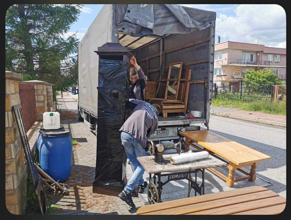 Dwóch mężczyzn ładuje owiniętą szafę do ciężarówki podczas przeprowadzki, w tle meble i maszyna do szycia.