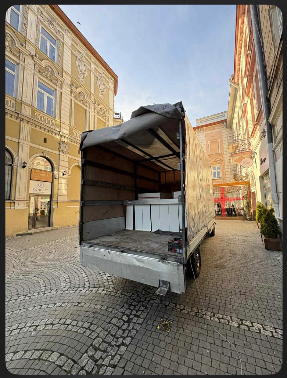 Otwarta paka busa z białymi kartonami na tle brukowanej uliczki i kamienic. Częściowo zwinięta plandeka. Widok z tyłu pojazdu.