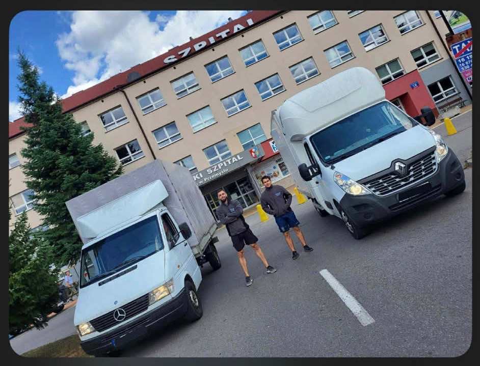 Dwa busy transportowe, Mercedes i Renault, zaparkowane przed budynkiem szpitala. Dwóch mężczyzn stoi na jezdni, prezentując pojazdy do transportu.