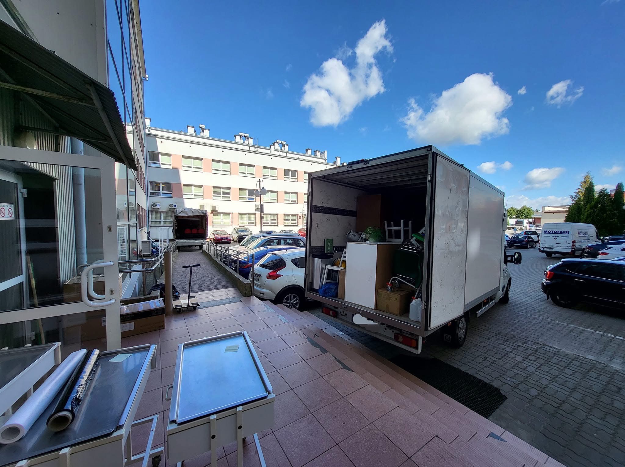 Otwarty furgon załadowany meblami i kartonami, zaparkowany przed budynkiem. Widoczne akcesoria do pakowania i transportu. Błękitne niebo z chmurami w tle.