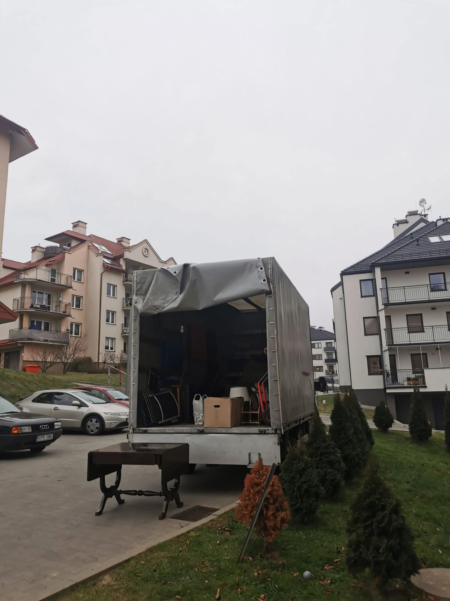 Otwarta paka samochodu dostawczego załadowana meblami i kartonami na tle budynków mieszkalnych. Na pierwszym planie stół z ciemnego drewna.