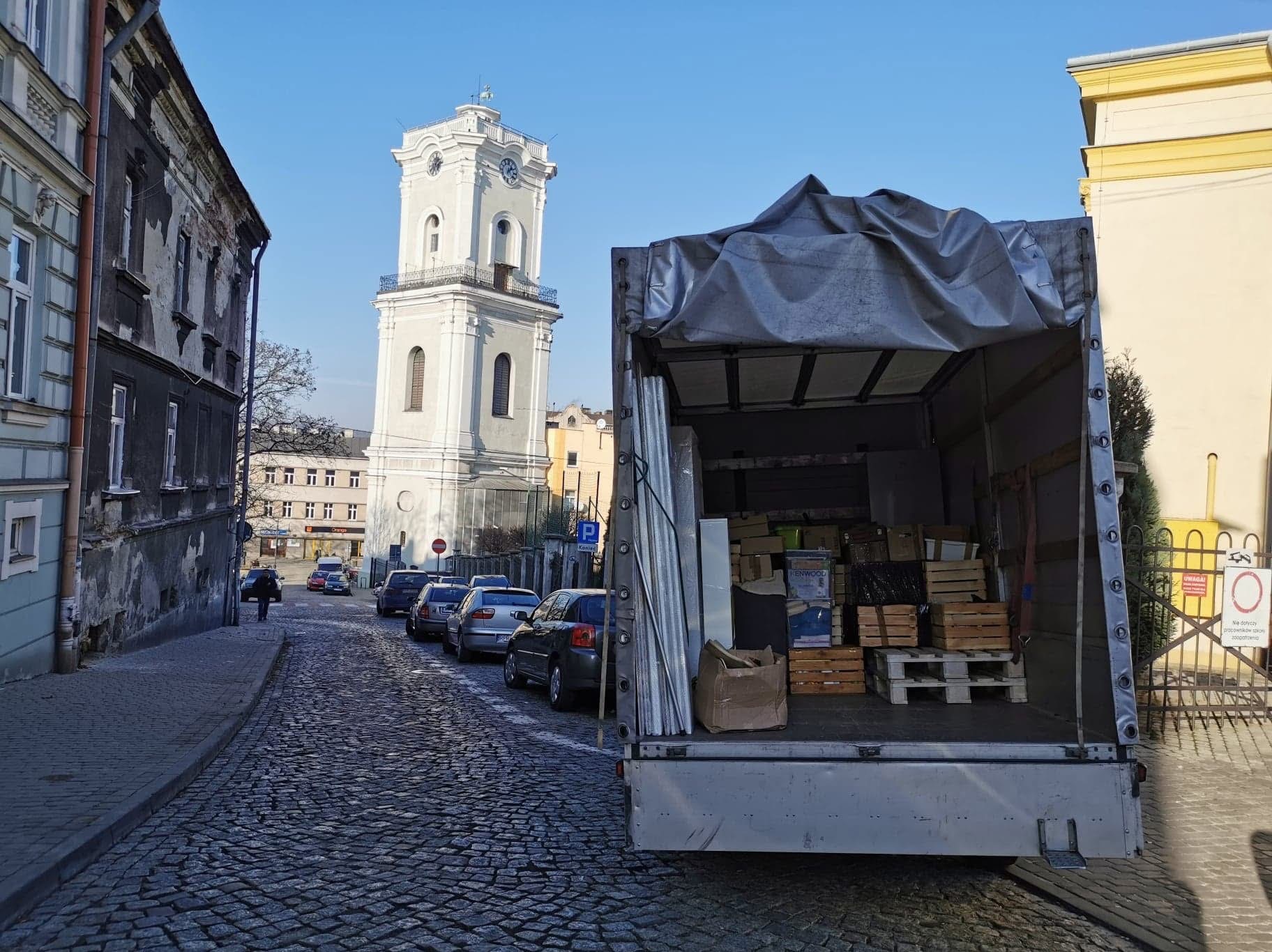 Otwarta paka busa wypełniona kartonami i skrzynkami na tle miejskiej uliczki z brukowaną nawierzchnią i wieżą zegarową w tle. Słoneczny dzień.