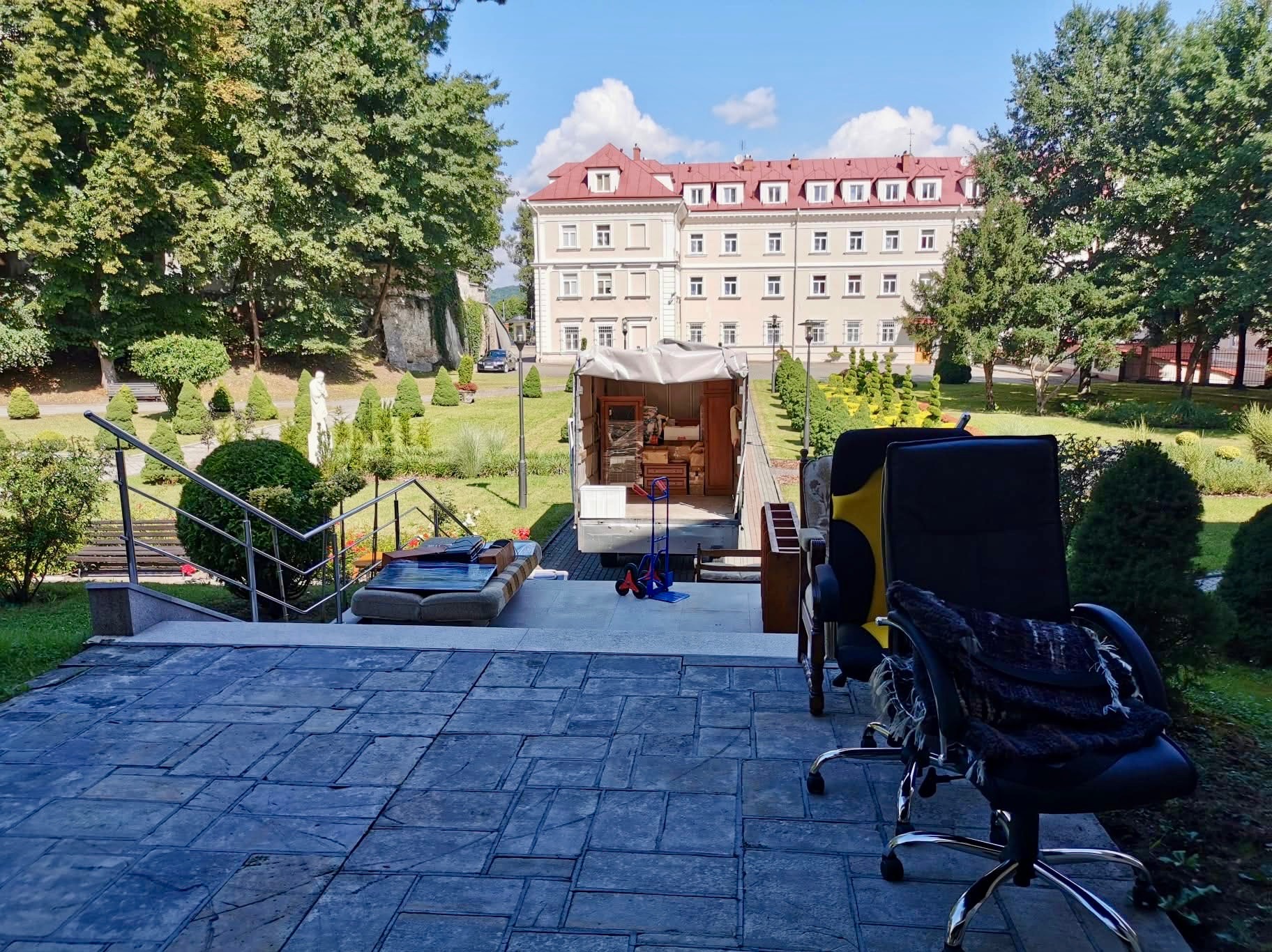 Przeprowadzka z eleganckiego budynku: meble biurowe i sofa na zewnątrz, w tle otwarty samochód dostawczy i budynek z czerwonym dachem w otoczeniu zieleni.