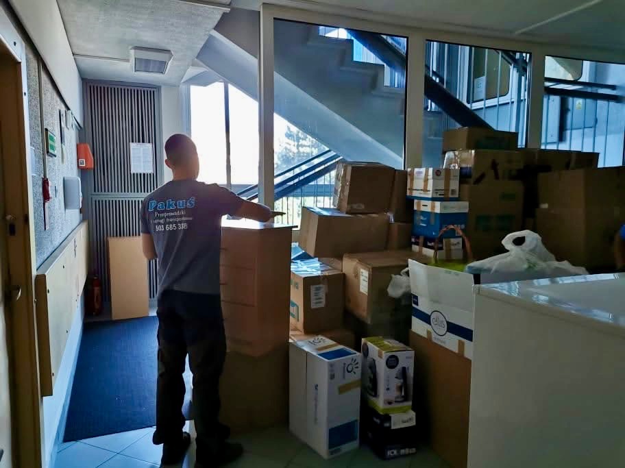 Pracownik firmy przeprowadzkowej układa kartony w korytarzu biurowca. Widoczne schody i okna. Na koszulce logo firmy.