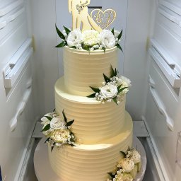 Iwona Korba - Trzypiętrowy tort weselny w chłodnym odcieniu bieli, zdobiony świeżymi białymi kwiatami i drewnianą ozdobą z sylwetkami pary młodej, umieszczony w chłodziarce.