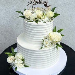 Iwona Korba - Dwupoziomowy tort weselny z białym kremem, zdobiony białymi różami i topperem 'Państwo Młodzi', na czarnym stole. Minimalistyczny design, elegancki wygląd.