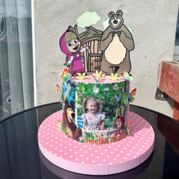 Iwona Korba - Tort urodzinowy z motywem Masha i Niedźwiedź, ozdobiony zdjęciem dziecka i motylami, na różowym podkładzie w białe kropki, stojący na czarnym stole.