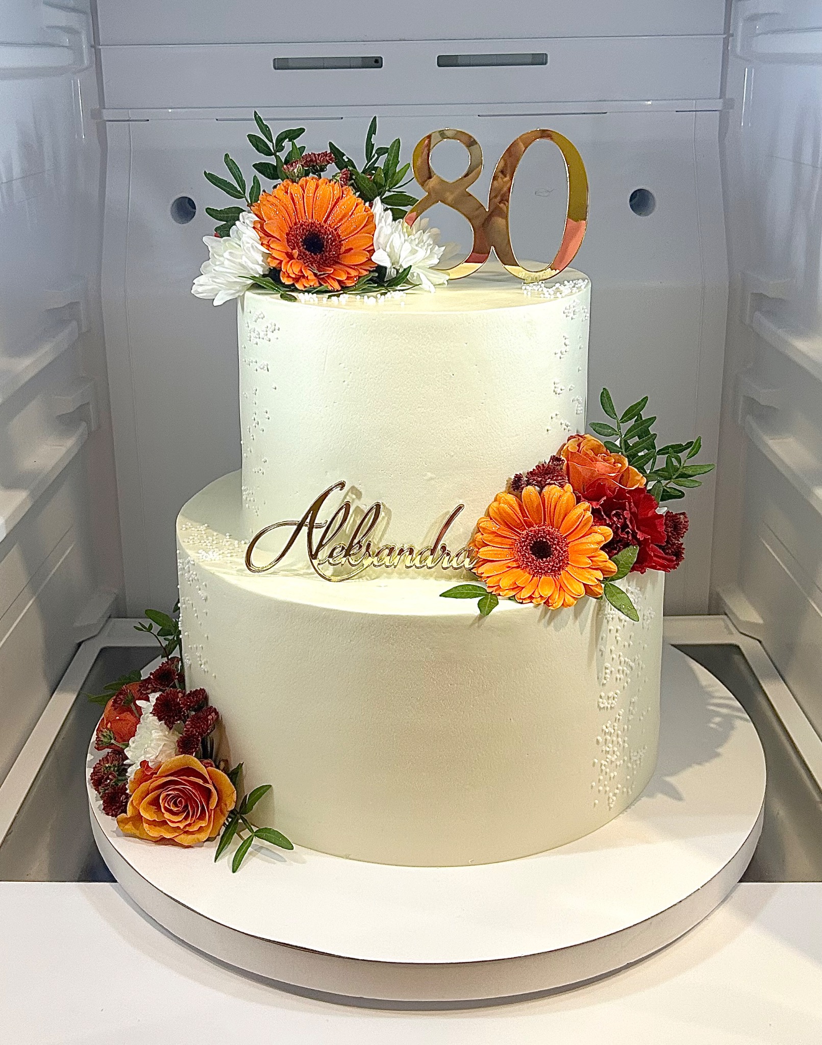 Dwupoziomowy tort urodzinowy z kremową polewą, kwiatami i złotym napisem 'Aleksandra' oraz '80', umieszczony w chłodziarce.