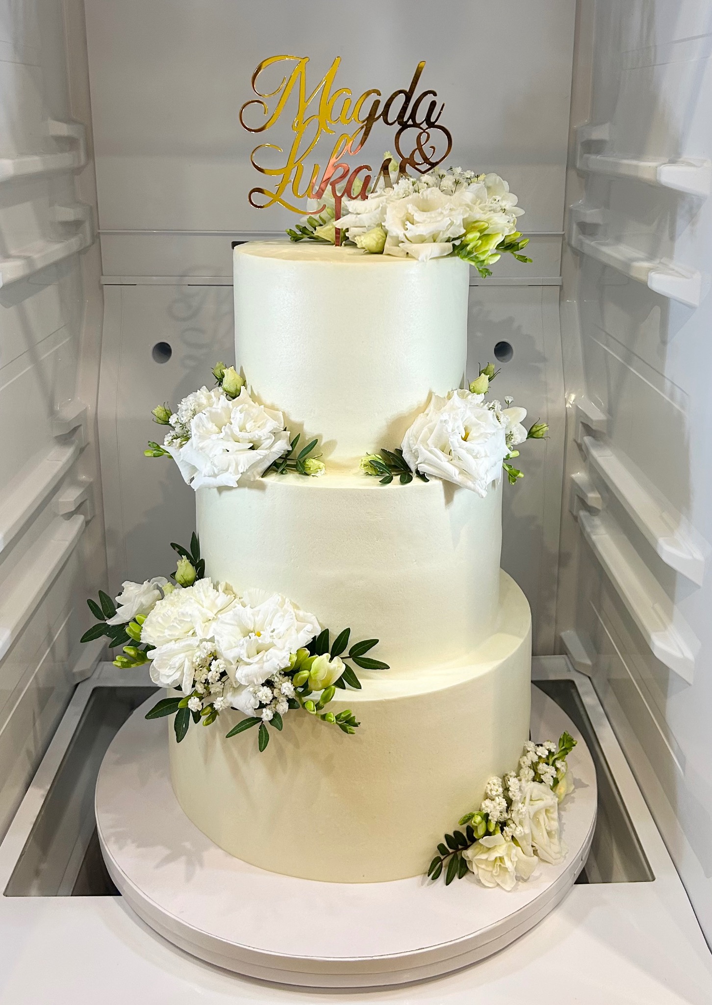 Elegancki, trzypiętrowy tort weselny w chłodnej tonacji bieli, zdobiony świeżymi, białymi kwiatami i złotym topperem z imionami 'Magda & Łukasz', umieszczony w chłodziarce.