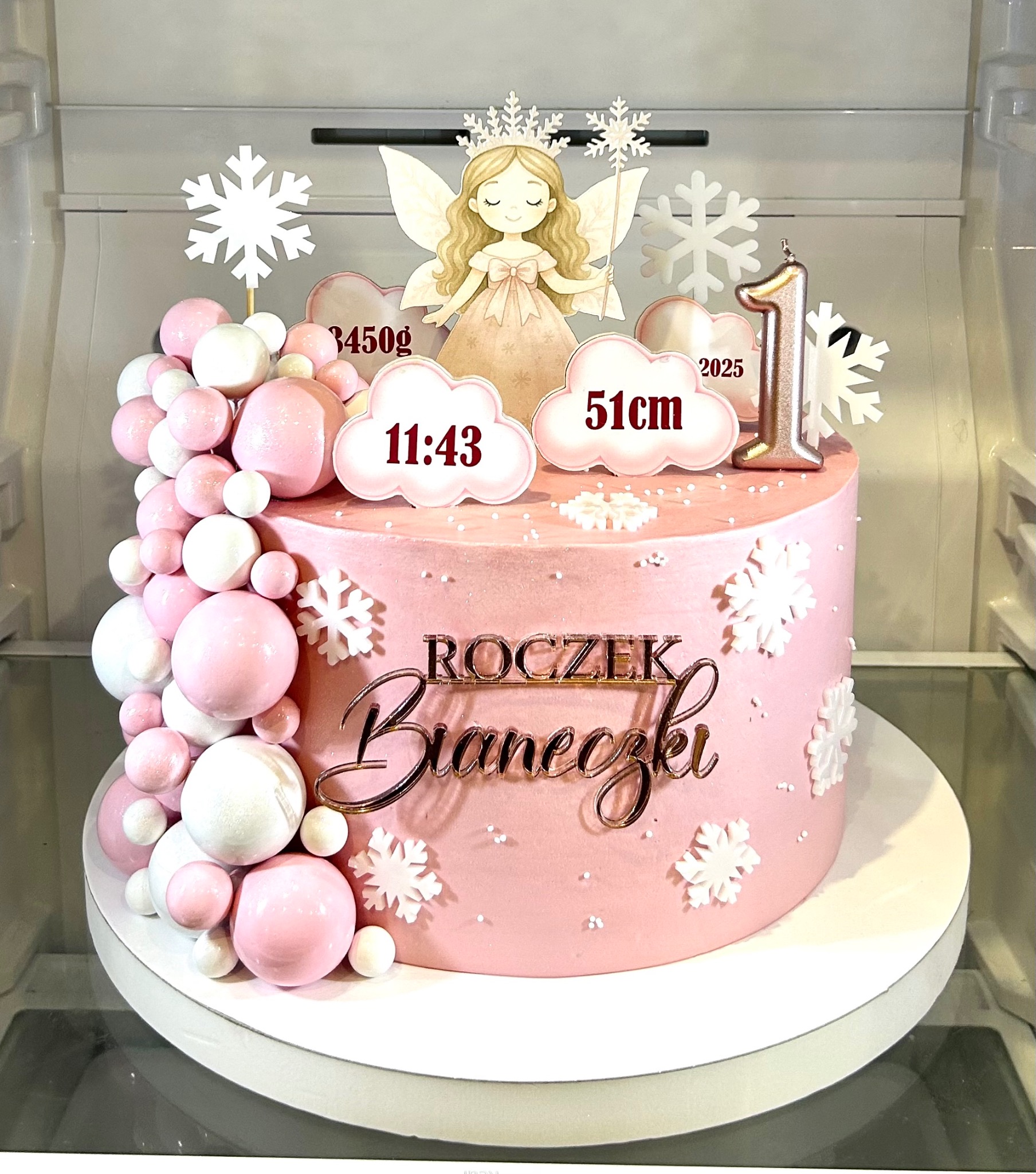 Różowy tort na roczek z dekoracjami: śnieżynki, figurka wróżki, balony, napisy z imieniem i datą urodzenia. Całość w chłodnej tonacji, w lodówce.