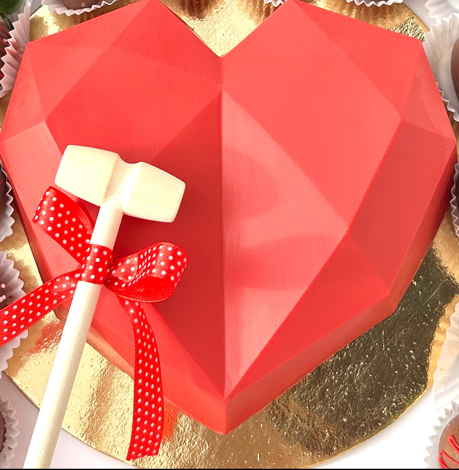 Czerwone, geometryczne serce z czekolady na złotym podkładzie, ozdobione młoteczkiem i czerwoną wstążką w białe kropki. Słodki upominek.
