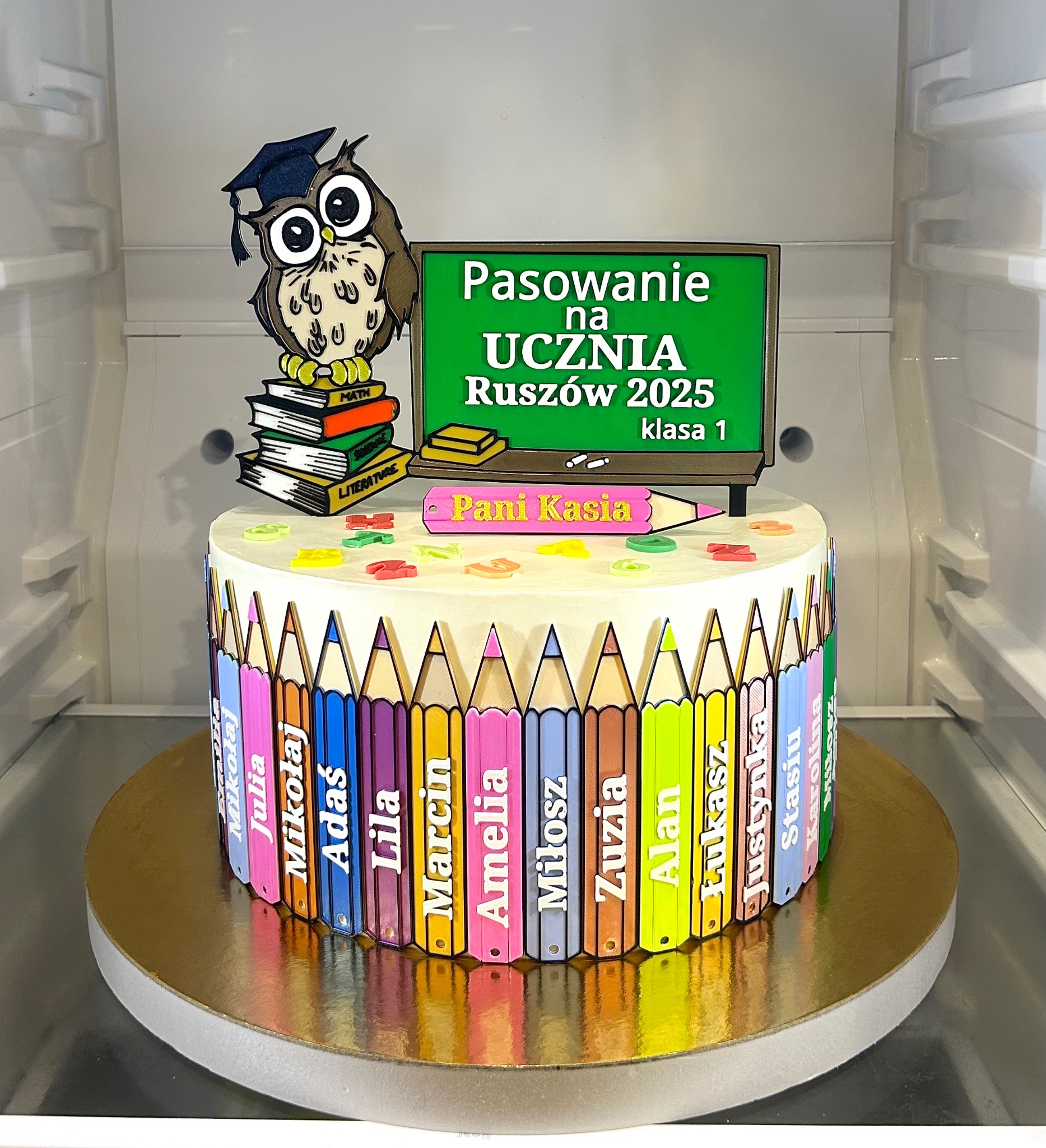 Tort na pasowanie ucznia z motywem kredek z imionami dzieci, sowy w birecie i napisem 'Pasowanie na Ucznia Ruszów 2025'. Dekoracja w chłodnej tonacji, w lodówce.