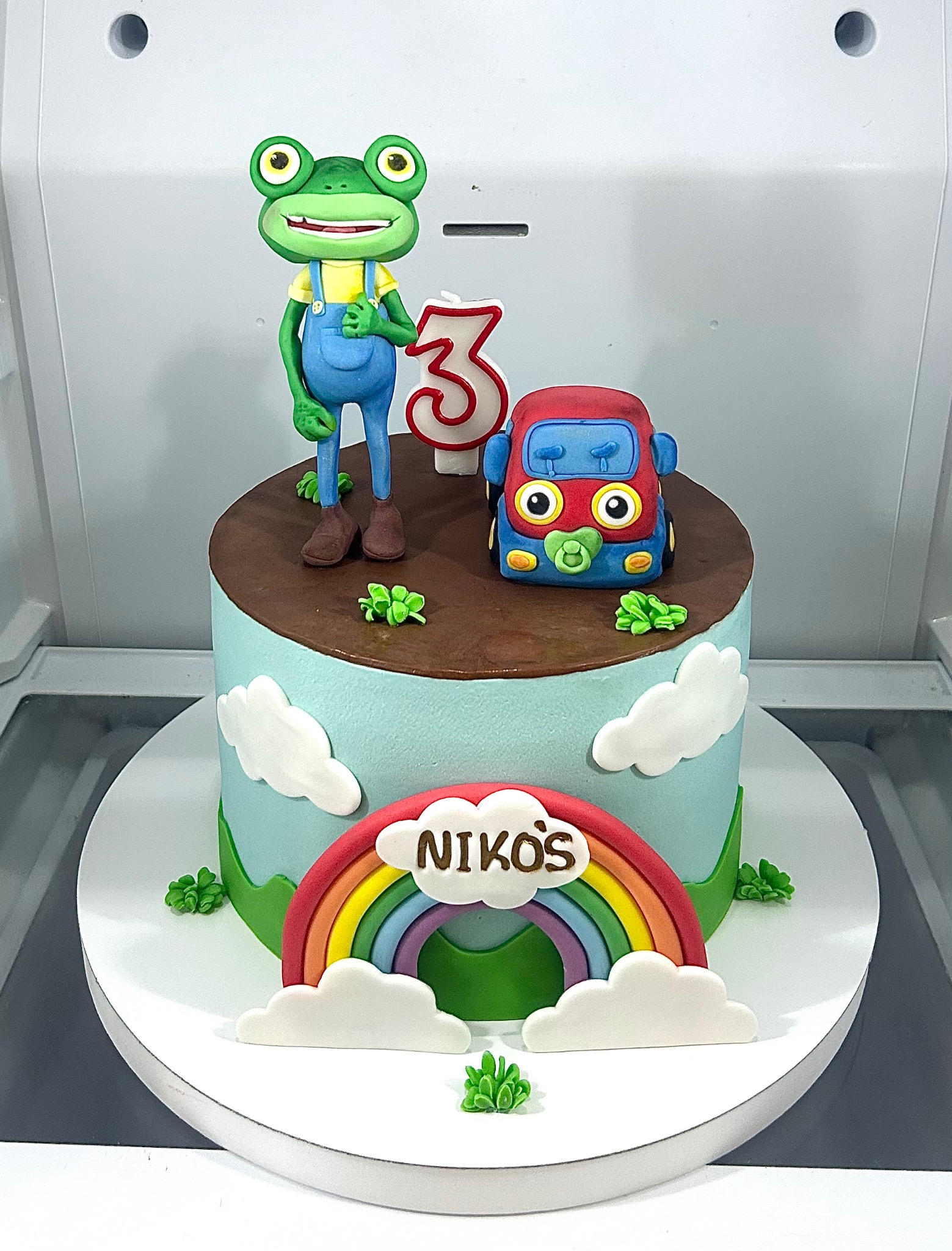 Udekorowany tort urodzinowy z figurką żaby, autka z smoczkiem i tęczą z napisem 'Nikoś', świeczka w kształcie cyfry 3. Pastelowe kolory, słodki motyw.