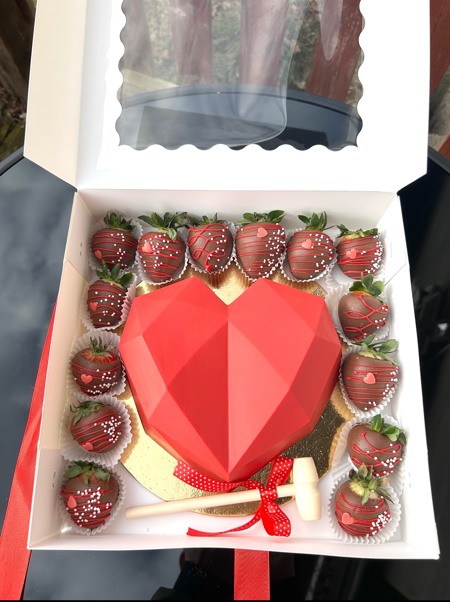 Otwarty box z czekoladowymi truskawkami i czerwonym sercem z czekolady, zdobionym wstążką. Dołączony mały młoteczek do rozbijania serca.