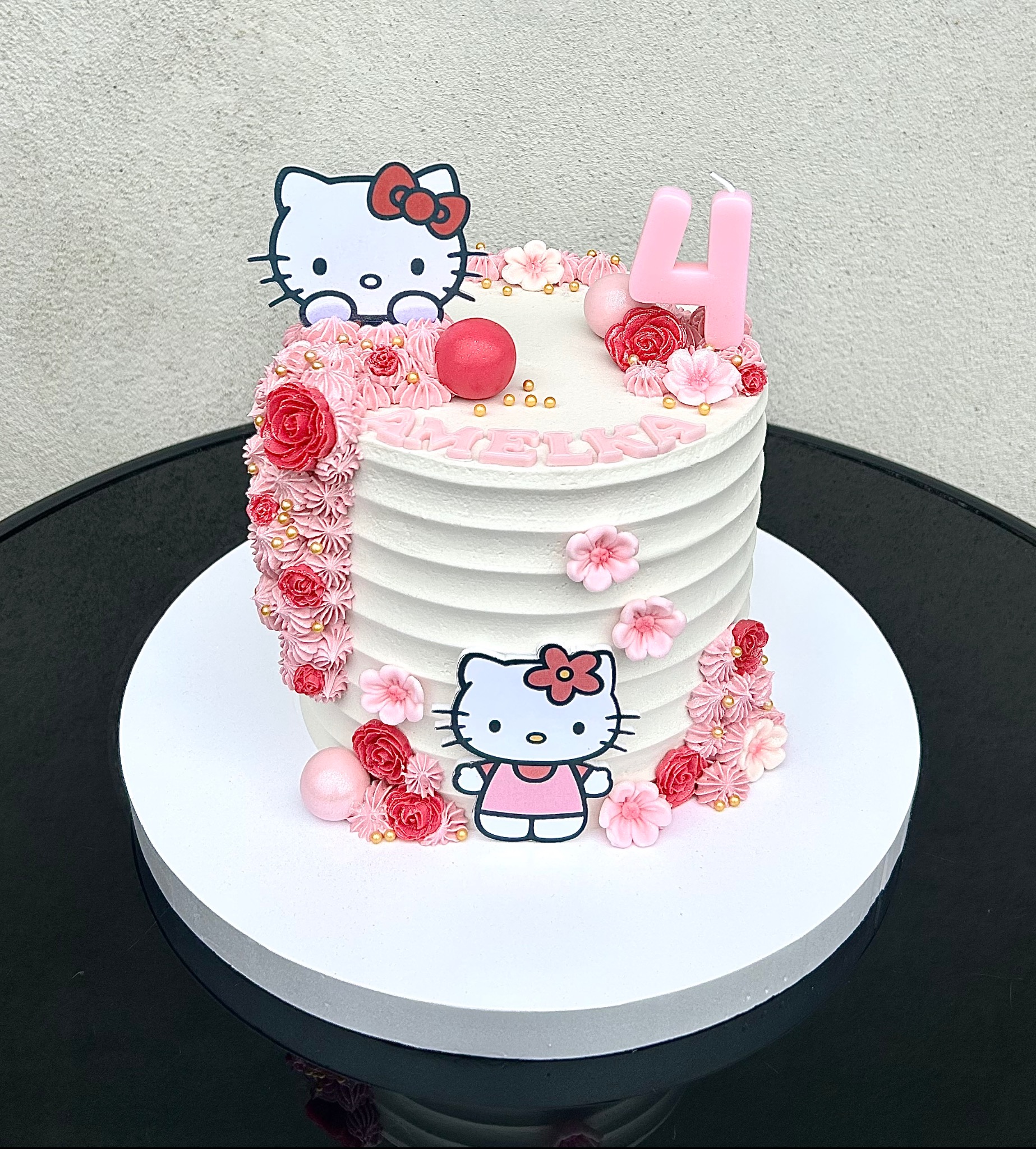 Tort urodzinowy dla dziewczynki z motywem Hello Kitty, zdobiony różowymi kwiatami, bezami i świeczką w kształcie cyfry 4. Napis 'Amelka' na torcie.