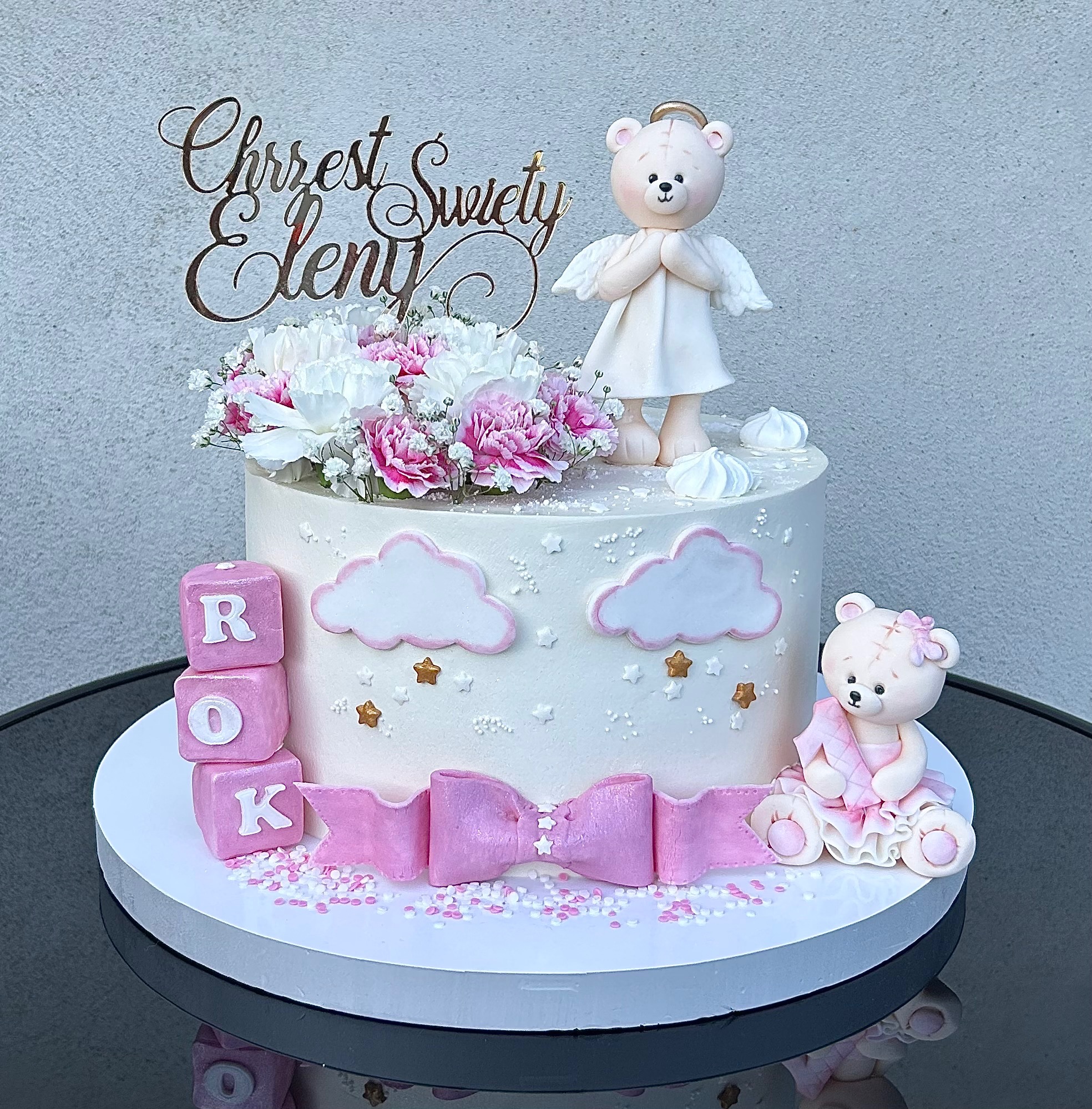 Tort okolicznościowy z dekoracjami: kwiaty, figurki misiów, kostki z literami ROK, różowa kokarda, chmurki i gwiazdki. Na torcie napis 'Chrzest Święty Eleny'.