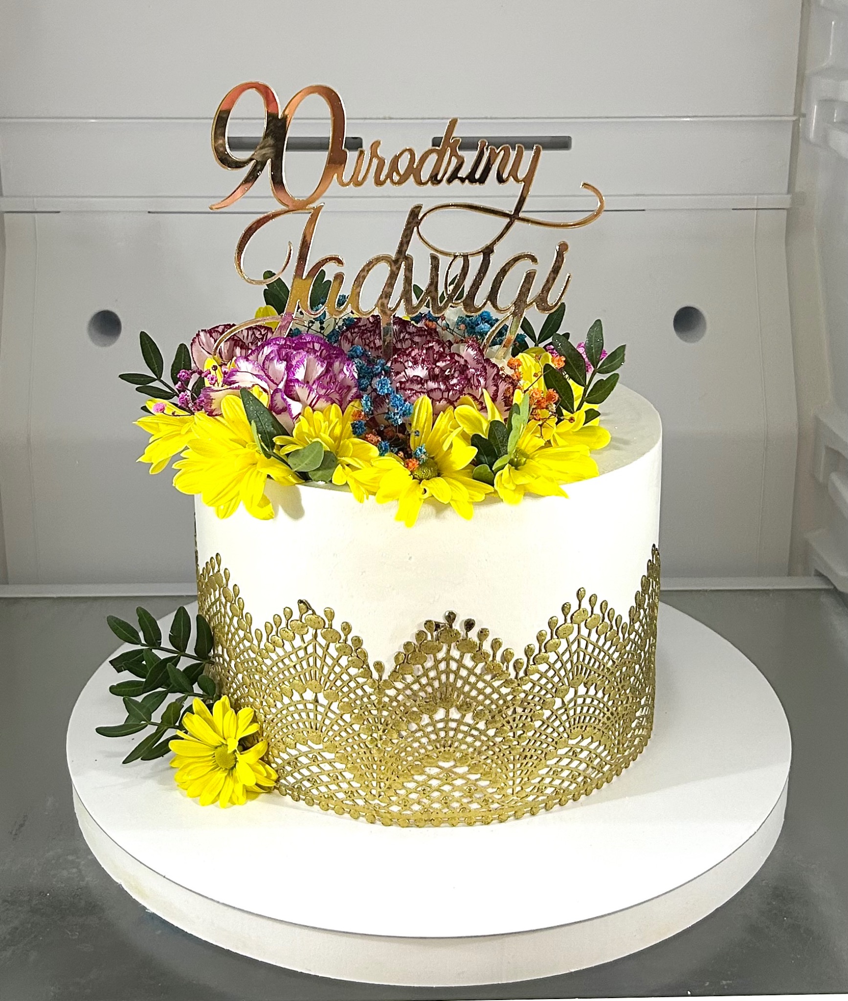 Tort urodzinowy z dekoracją kwiatową i złotym napisem '90 Urodziny Jadwigi', umieszczony na białym podeście.