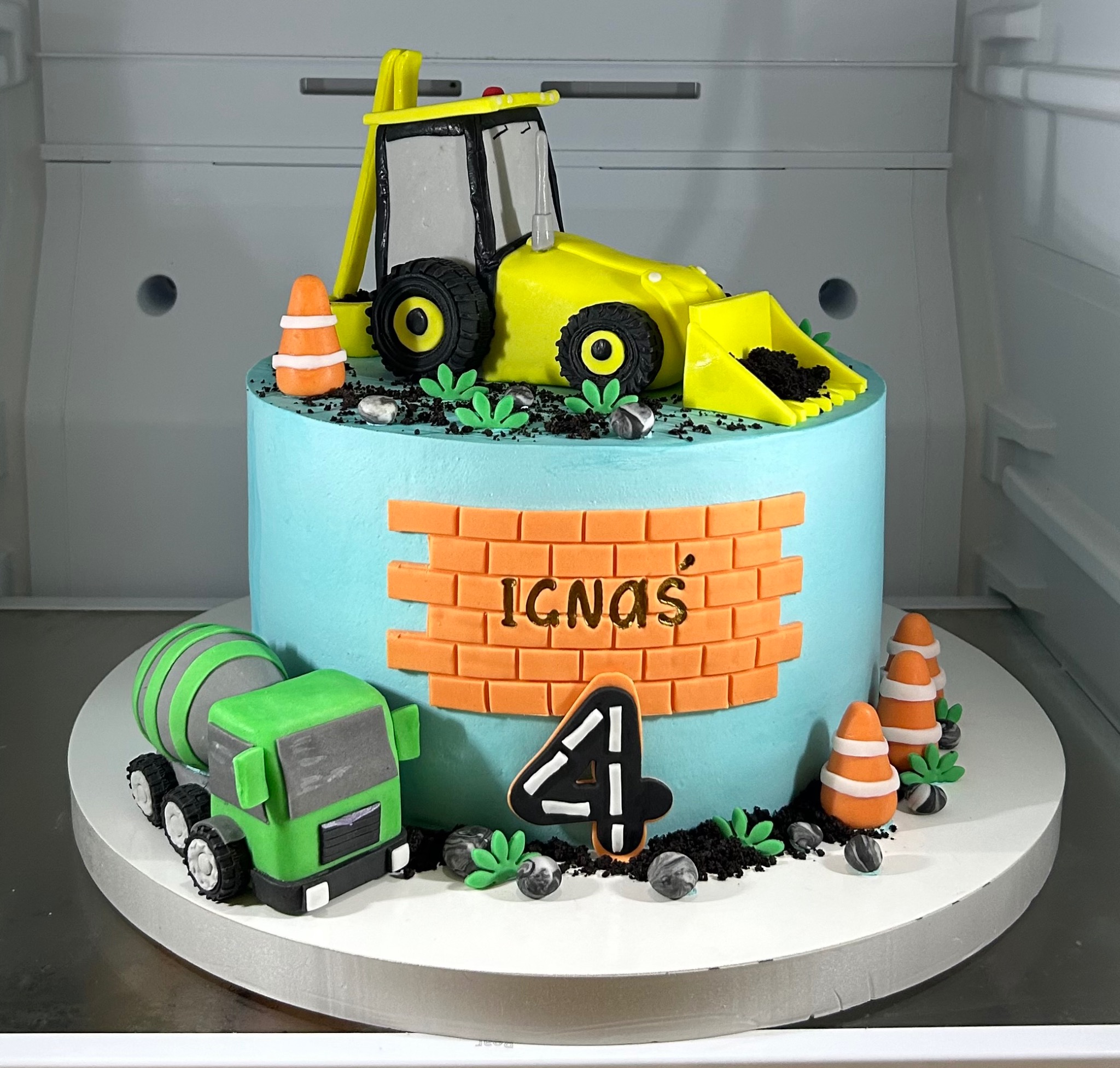 Urodzinowy tort w stylu budowlanym: błękitny tort z dekoracjami z lukru - koparka, betoniarka, ceglany mur z imieniem 'Ignas' i cyfra 4. Całość umieszczona w chłodziarce.