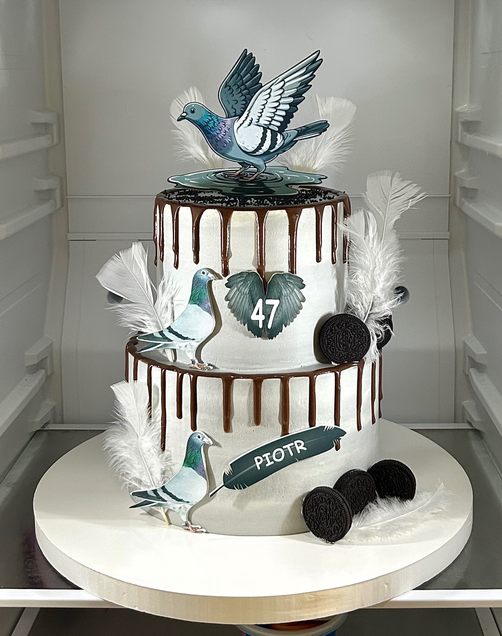 Dwupiętrowy tort z białym kremem, czekoladowym dripem, dekoracjami z piór, ciastek Oreo i gołębi. Na torcie imię 'Piotr' i liczba '47'.