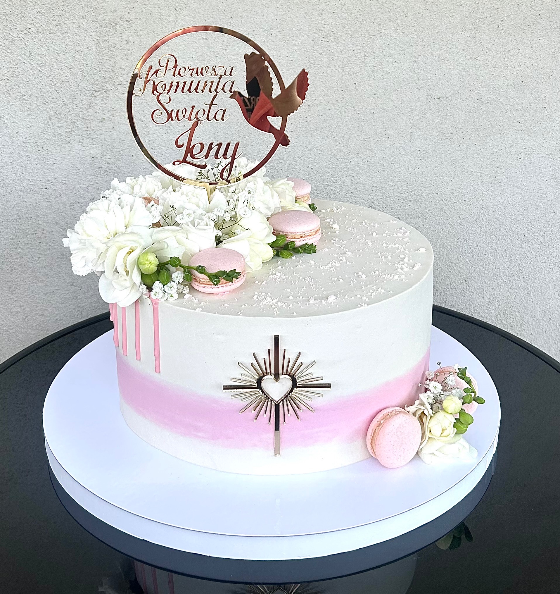 Tort komunijny z białym kremem, różowym ombre, dekoracjami z kwiatów, makaroników i topperem z napisem 'Pierwsza Komunia Święta Leny' oraz symbolem gołębicy.