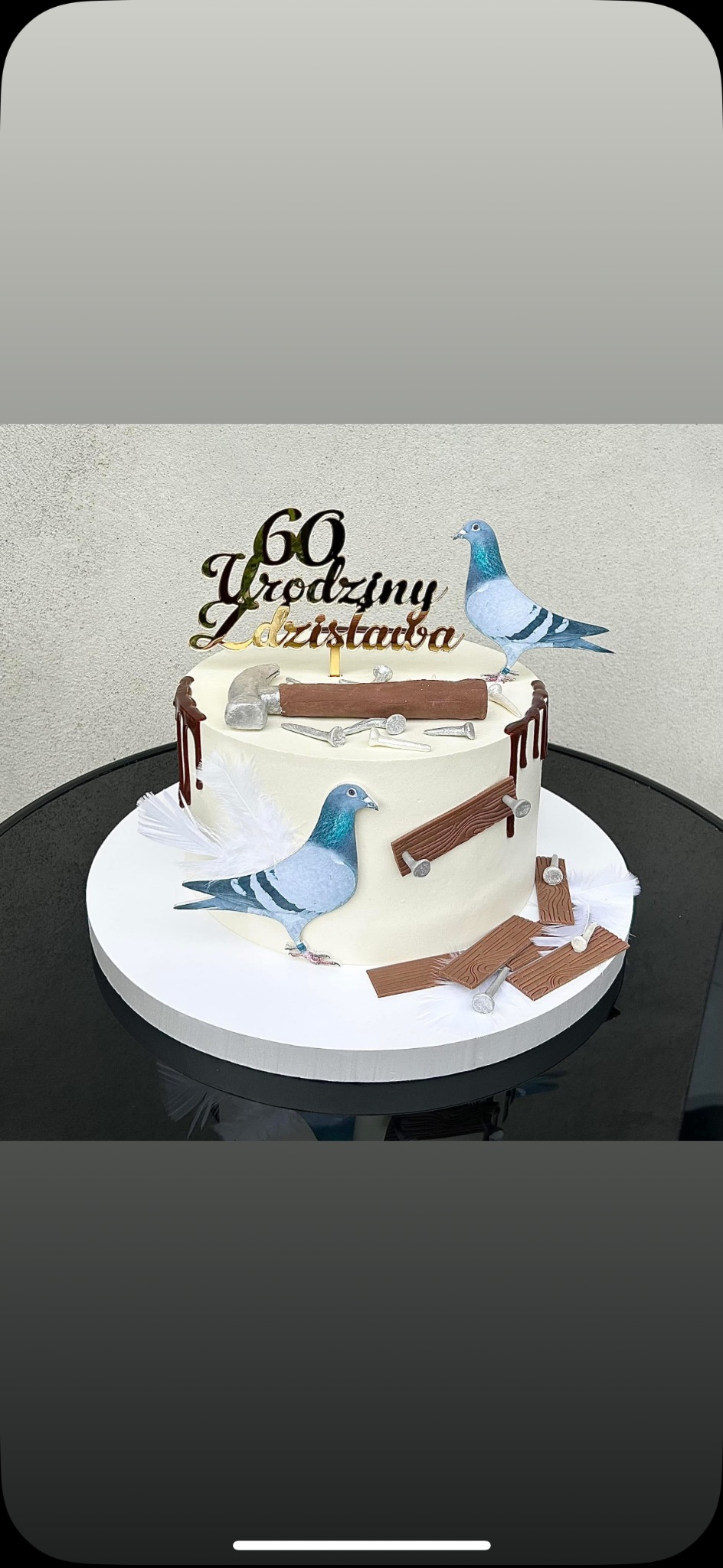 Tort urodzinowy z dekoracjami tematycznymi: gołębie, narzędzia stolarskie i napis '60 Urodziny Zdzisława'. Kremowy tort na czarnym stole.