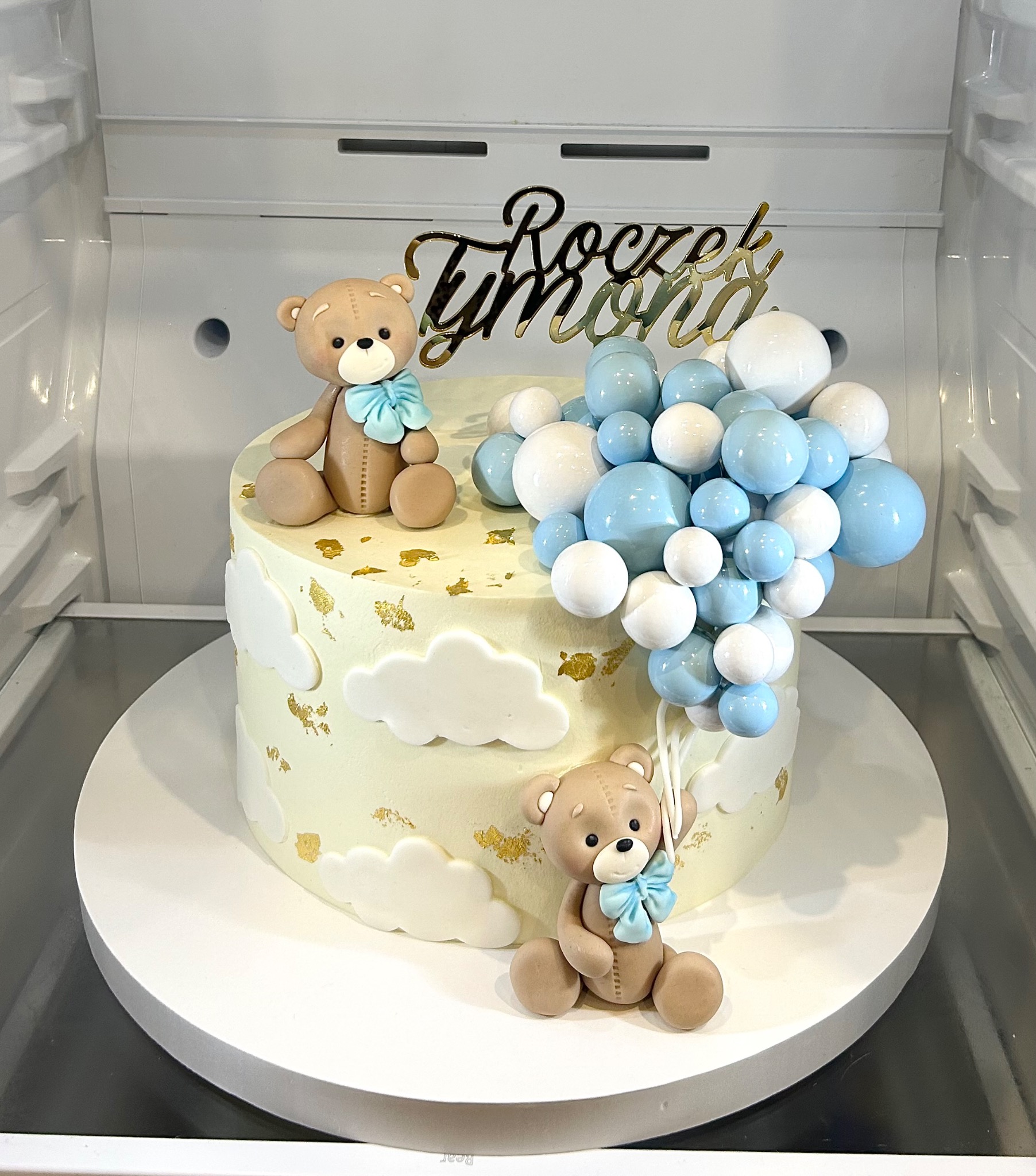 Tort na roczek z dekoracjami: dwa misie z kokardkami, biało-błękitne balony i złoty napis 'Roczek Tymona'. Kremowy tort z motywem chmur i złotych płatków.
